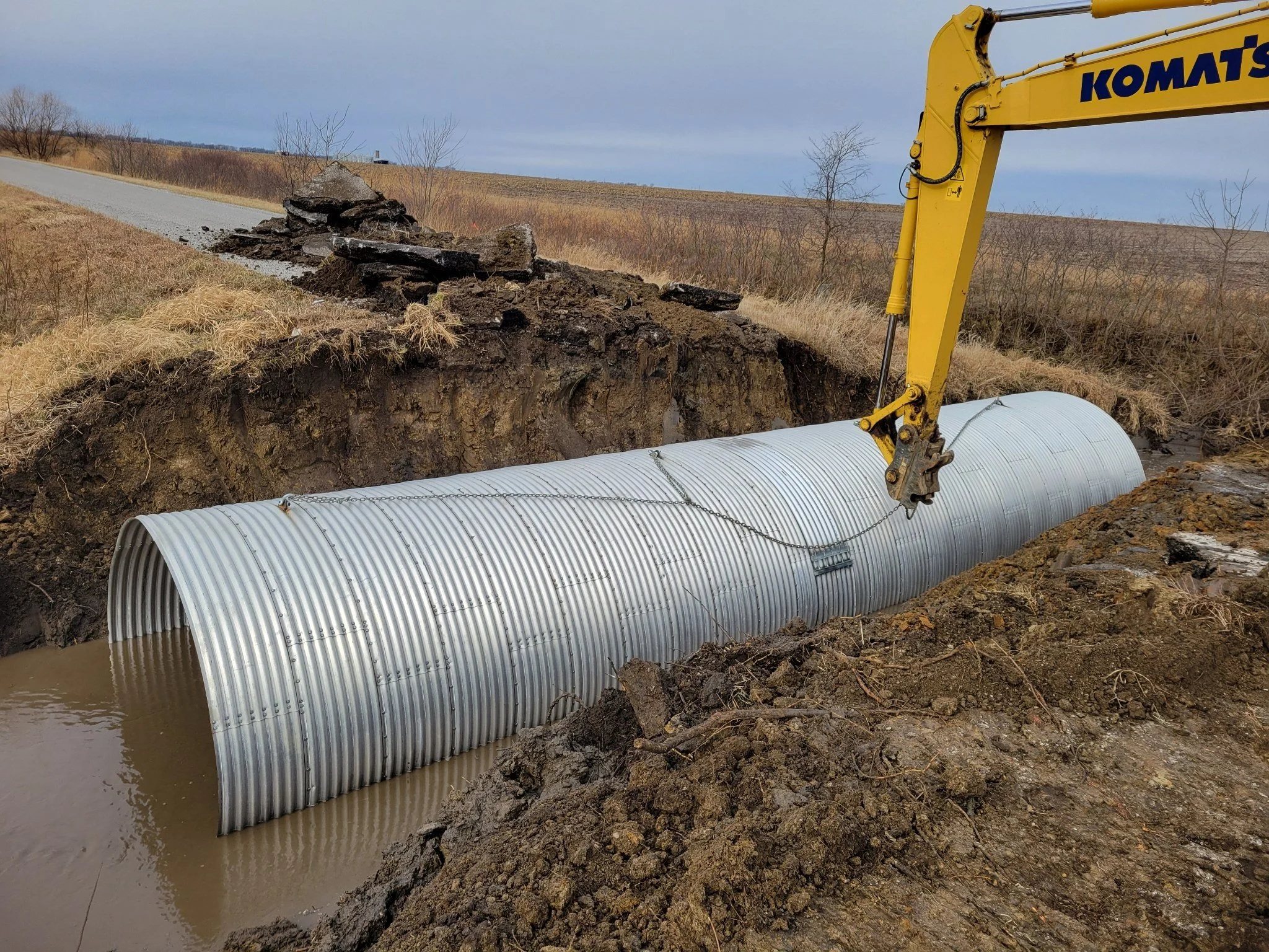 Construction site with a large corrugated metal pipe being installed in a trench by a yellow excavator.