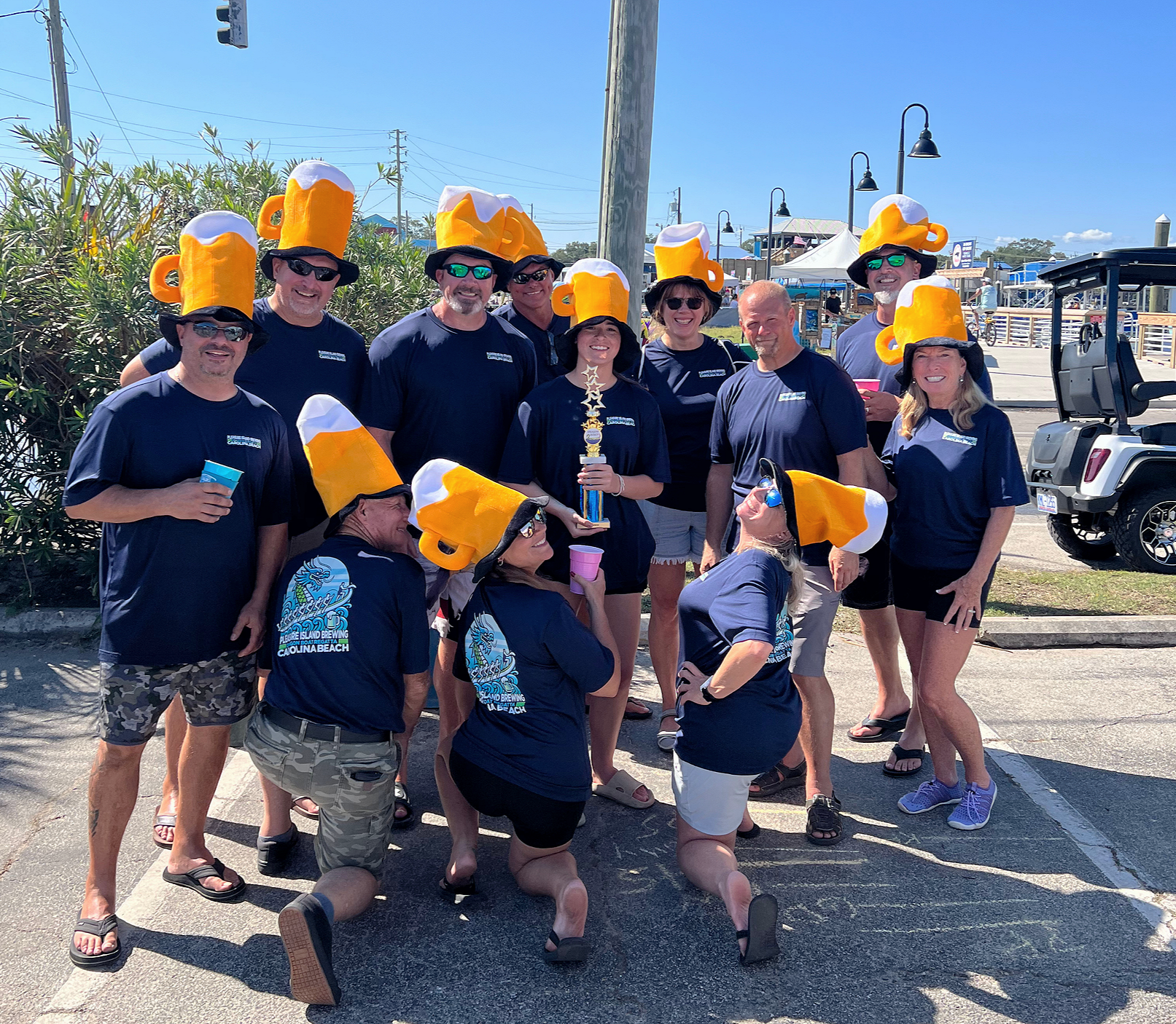 Group of people wearing beer mug hats, holding trophies and drinks, outdoors on a sunny day, likely celebrating a brewery event.