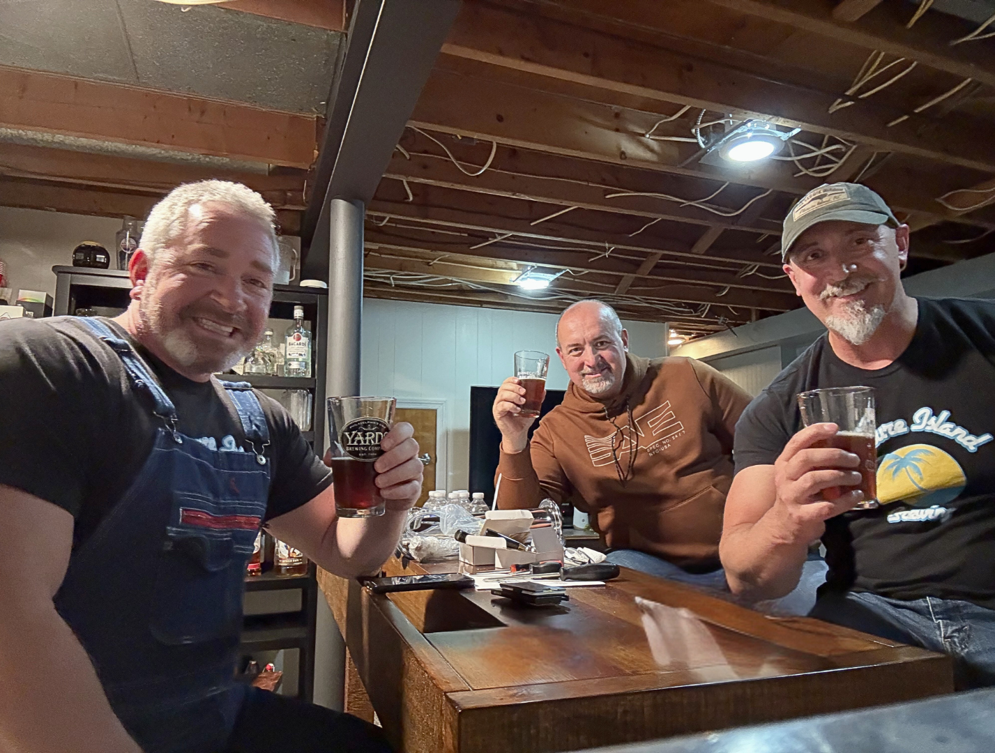 Three middle-aged men sitting at a bar, smiling and holding glasses of dark beer. The bar has a wooden top, and the background shows shelves with bottles of alcohol and bar equipment. The ceiling is exposed with visible wooden beams and wiring.