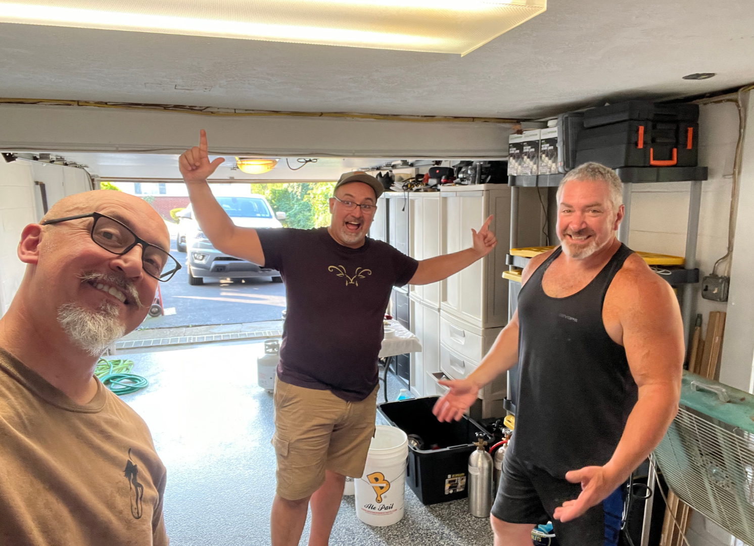 Three men in a garage, smiling and celebrating. One man on the left takes a selfie, the man in the middle has arms raised, and the man on the right gestures with open hands. Parking lot with vehicles visible outside.