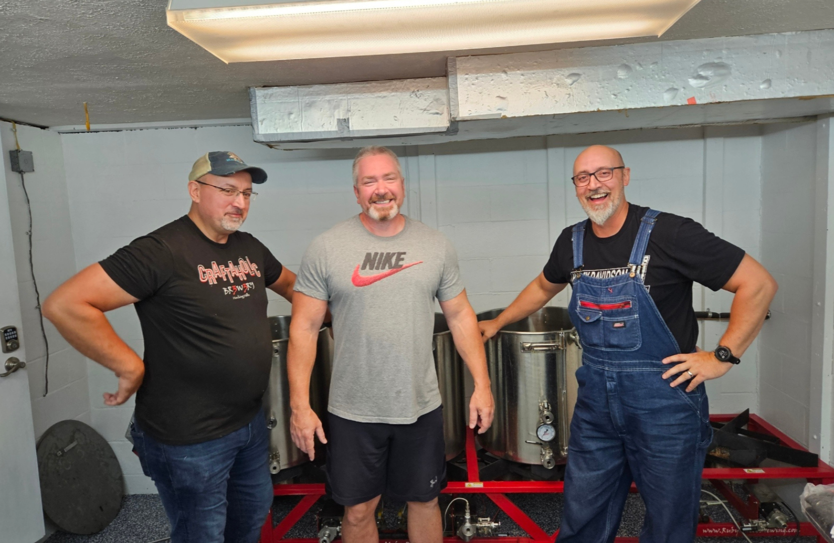 Three men standing together in a basement, smiling, with brewing equipment behind them. The man on the left wears glasses, a cap, and a black T-shirt. The man in the middle wears a gray Nike shirt. The man on the right wears glasses, a black T-shirt,