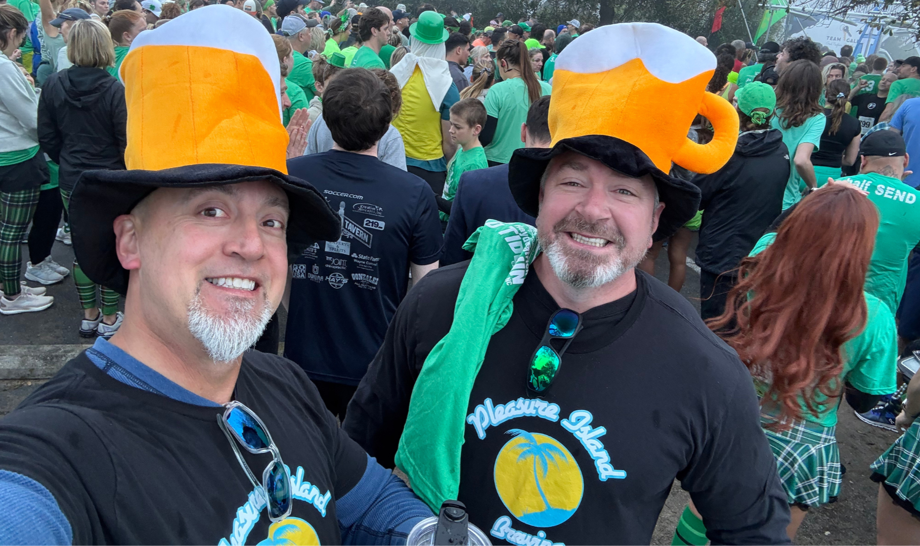Two men wearing large beer mug costumes as hats at a crowded outdoor event, with many people in green shirts around them.
