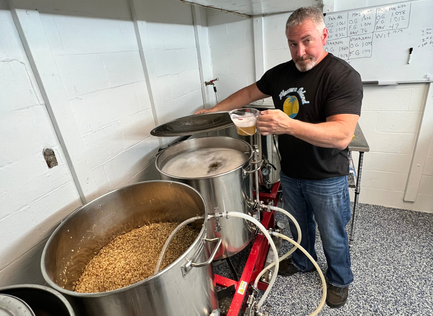 A man with gray hair and beard, wearing a black t-shirt with a pineapples design, stands in a brewery or brewing area, holding a glass of beer. He is pouring or about to pour the beer from a brewing vessel. Several large brewing tanks are positioned 