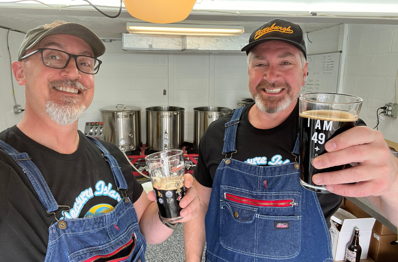 Two smiling men in black t-shirts and blue aprons holding glasses of dark beer, standing in front of brewing equipment in a brewery.
