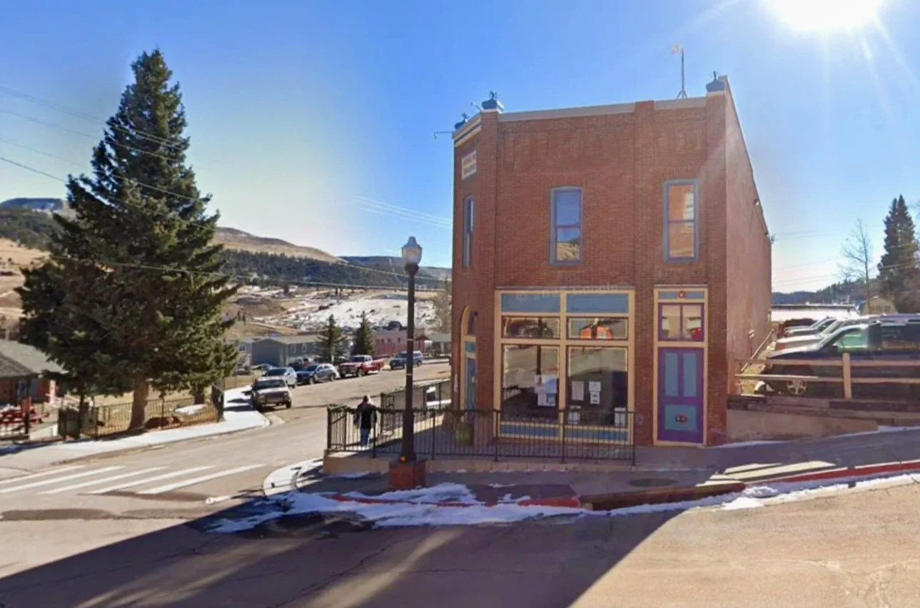 Front of the Aspen Mine Center West building that houses A Willow Bends Counseling Services in Cripple Creek, Colorado. A two-story brick building on the corner of the street with a lamp post and sidewalk railing in front.