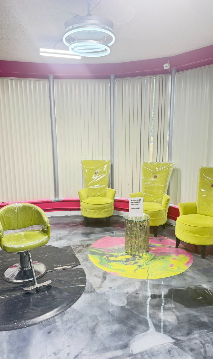 A waiting area with four lime green chairs, two of which are high-backed with plastic covers, and one lime green swivel chair. There is a colorful round rug with pink, yellow, and green paint splatters, and a small silver side table with a sign on it