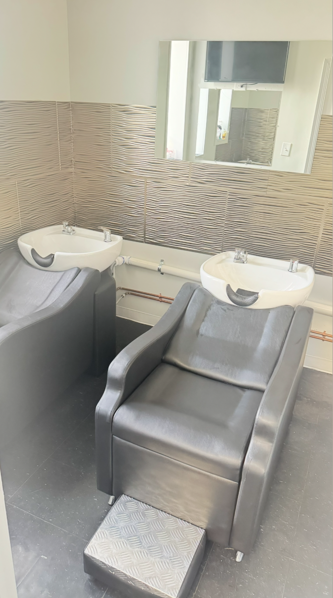 A salon chair with a hair wash basin behind it, set against a tiled wall with a mirror reflecting part of the room.