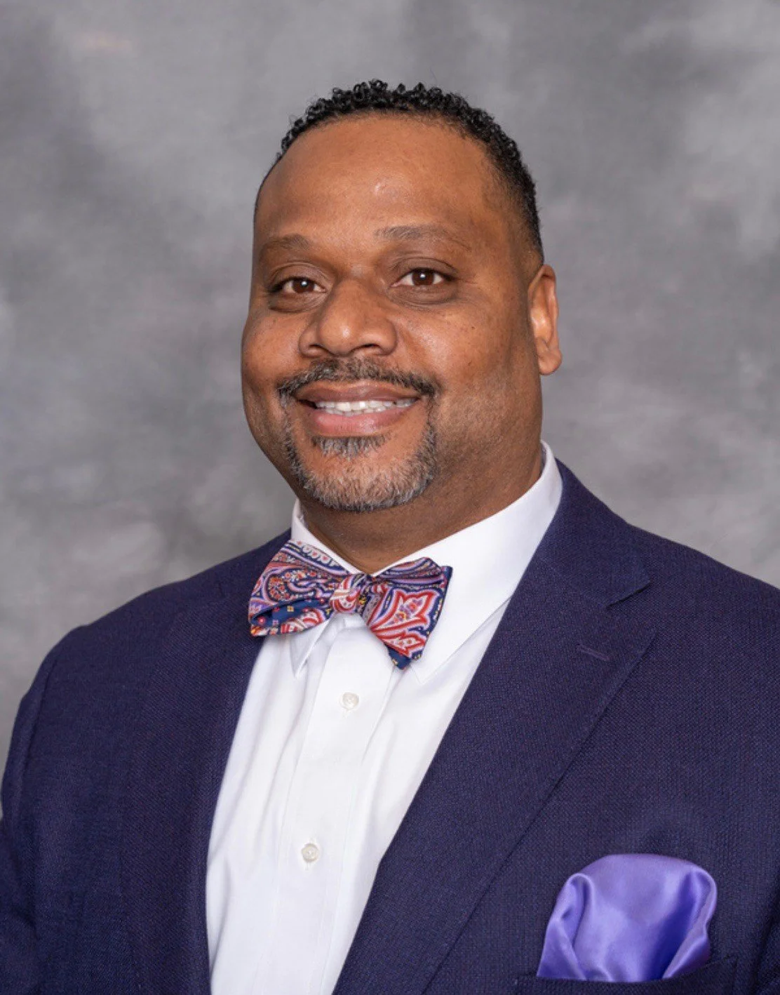 A professional portrait of a smiling man wearing a navy blue suit, a white shirt, a colorful patterned bow tie, and a purple pocket square, set against a gray background.