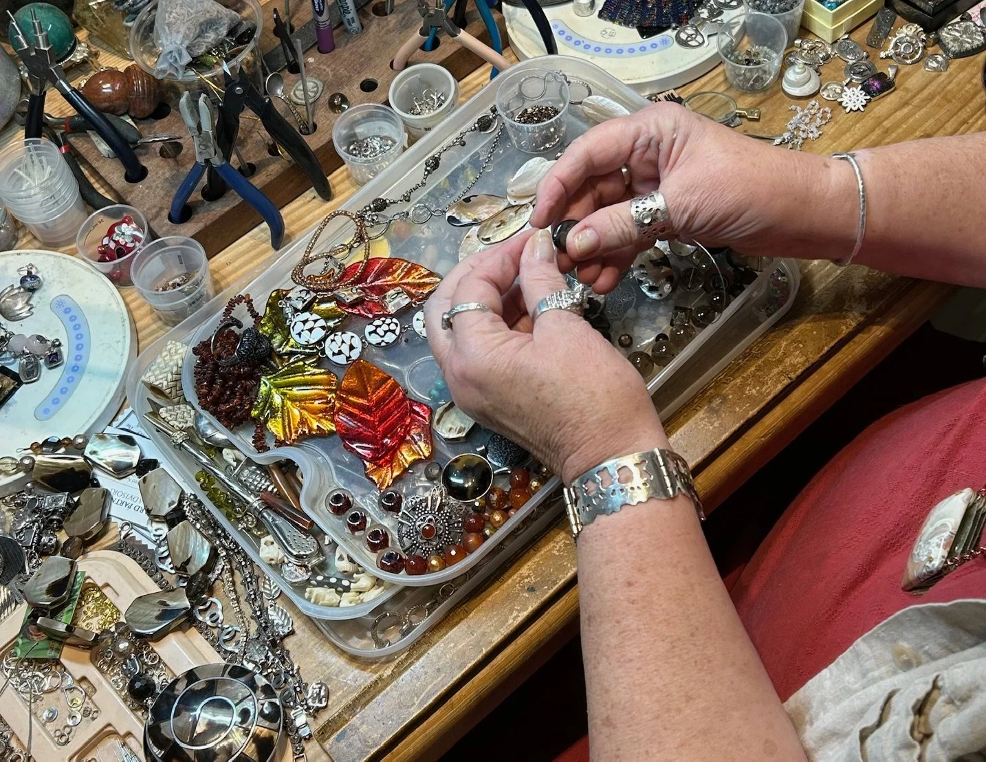 Kate Stockman in her studio working on jewelry assembly at a jeweler's workbench with various jewelry pieces, tools, and containers of beads and findings.