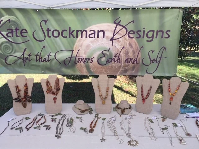 Jewelry display with necklaces and bracelets at an outdoor craft fair under a green banner that reads "Kate Stockman Designs, Art that Honors Earth and Self."
