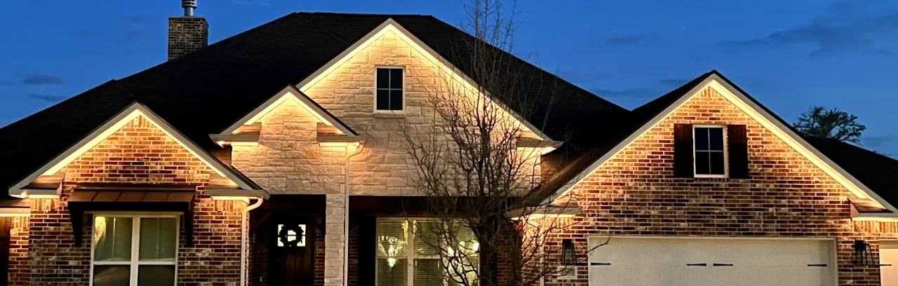 A two-story brick house with an illuminated front exterior at dusk, featuring a gabled roof, a garage, and windows with shutters.