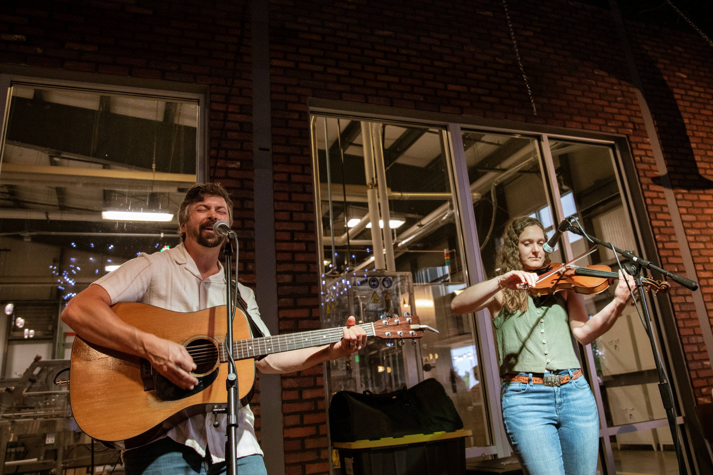 A man playing an acoustic guitar and singing into a microphone, and a woman playing a fiddle into a microphone, performing in an indoor venue with brick walls and large windows.