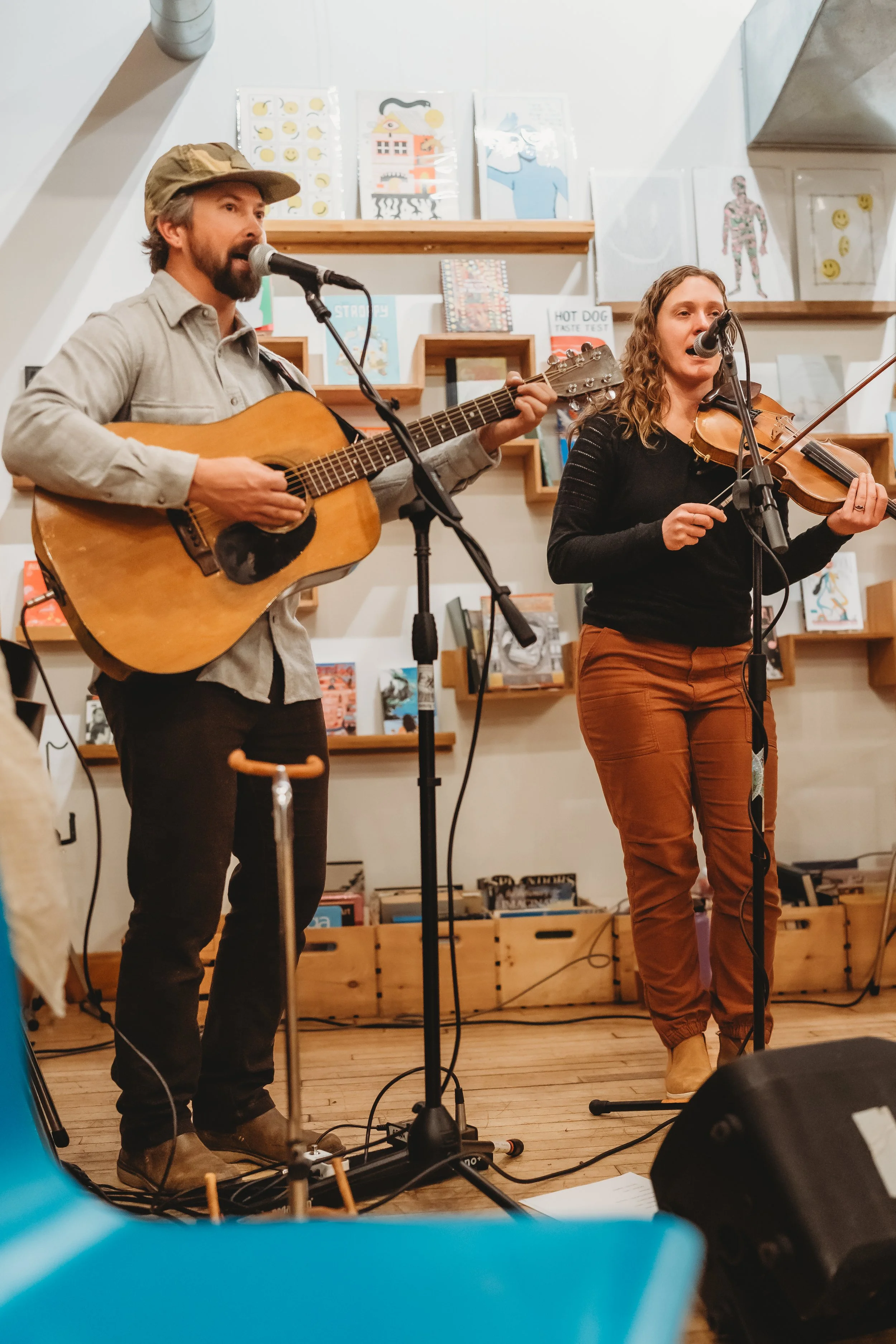 A man and woman performing musical act with microphone and instruments, in a cozy indoor space with books and artwork on the shelves.