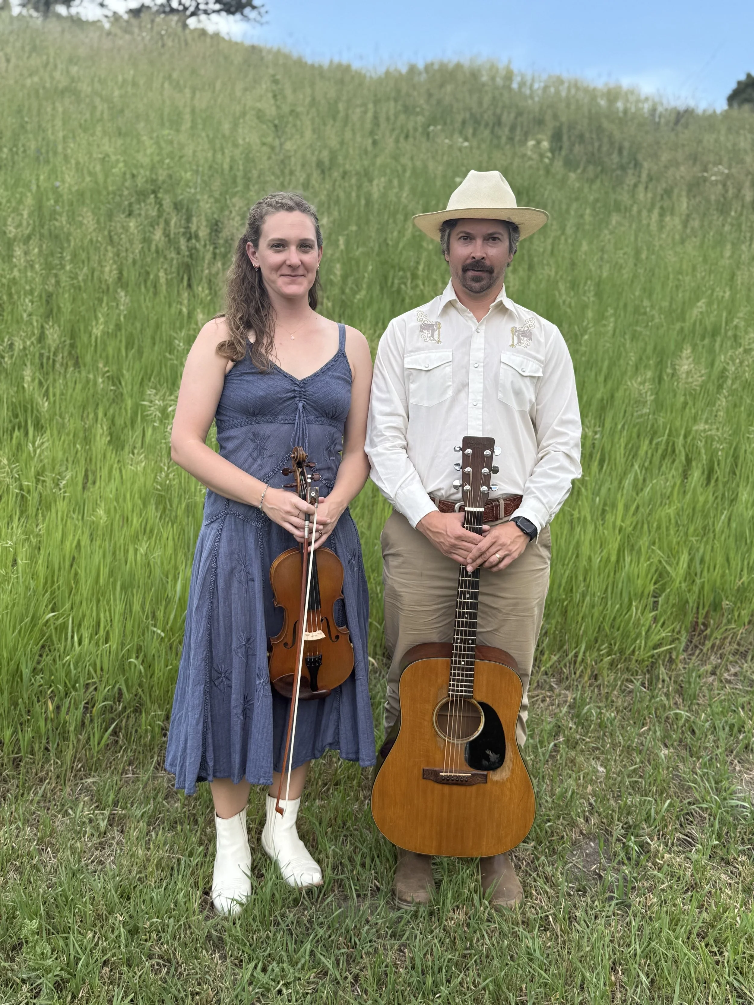 A woman holding a fiddle and a man holding a guitar standing together outdoors in a grassy field with a hill behind them, both dressed in casual, country-style clothing.