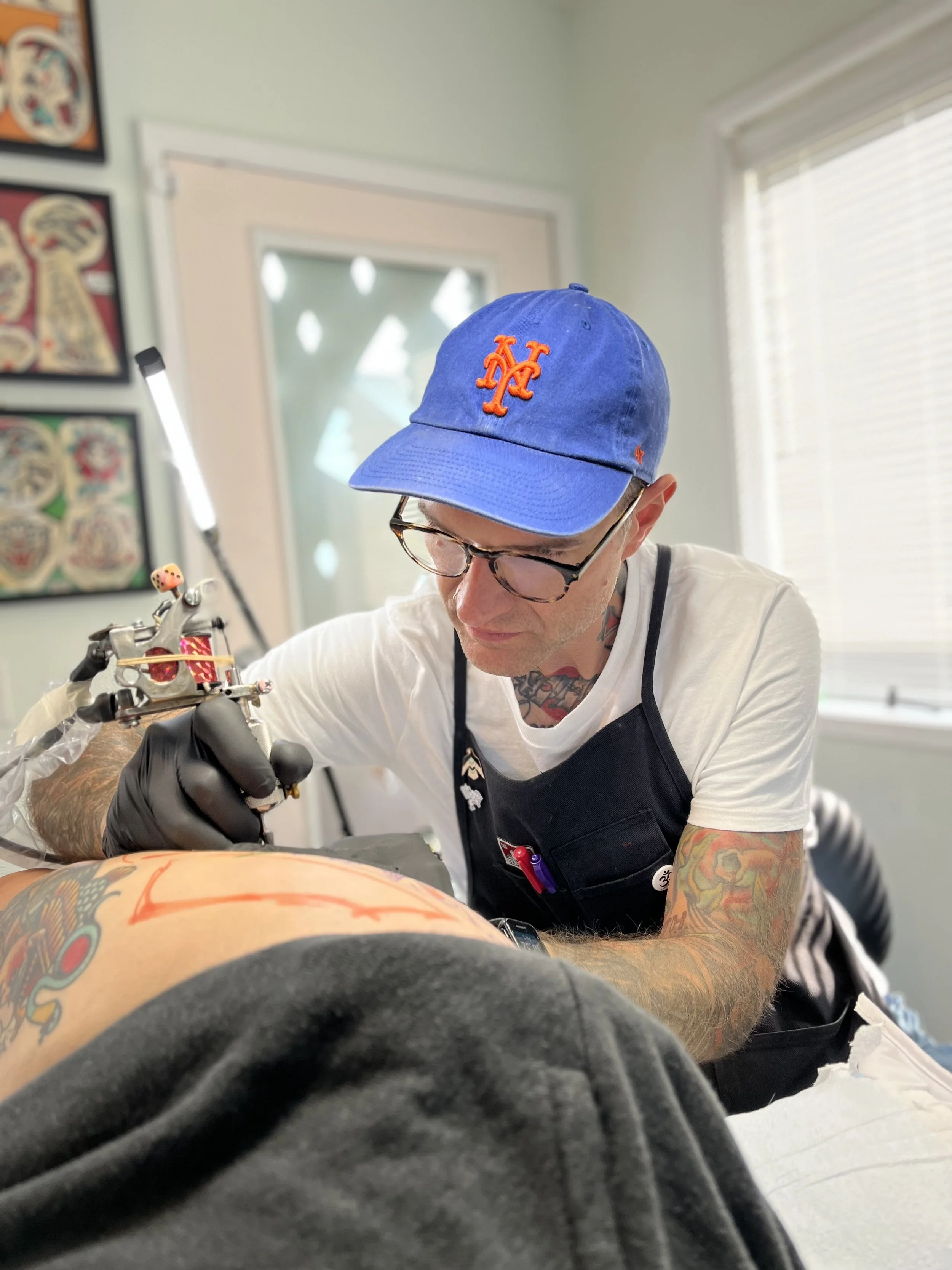Tattoo artist in a blue cap, glasses, and black apron working on a tattoo in a tattoo studio.