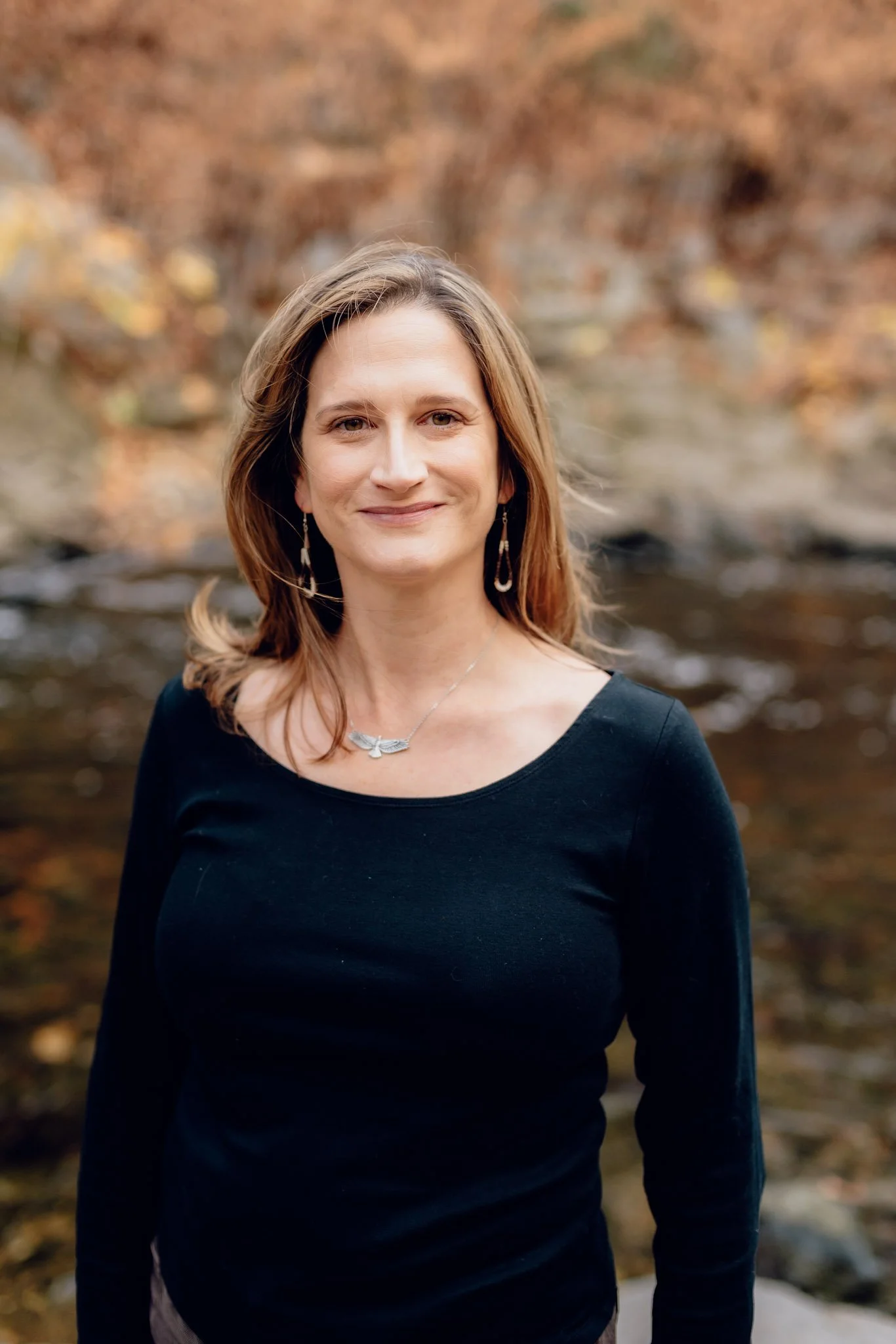 A woman with shoulder-length brown hair and light skin, wearing a black long-sleeve top and silver jewelry, standing outdoors in front of a blurred background of autumn trees and water.