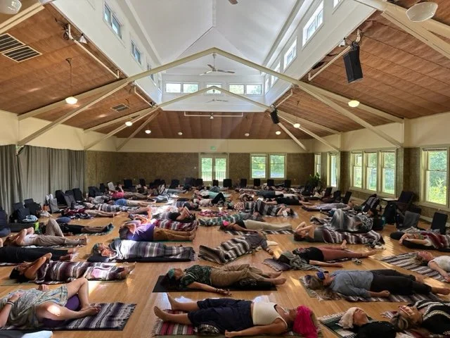 A large group of people lying down on mats in a spacious room with high vaulted ceiling, large windows, and wooden interior, participating in a yoga or meditation session.