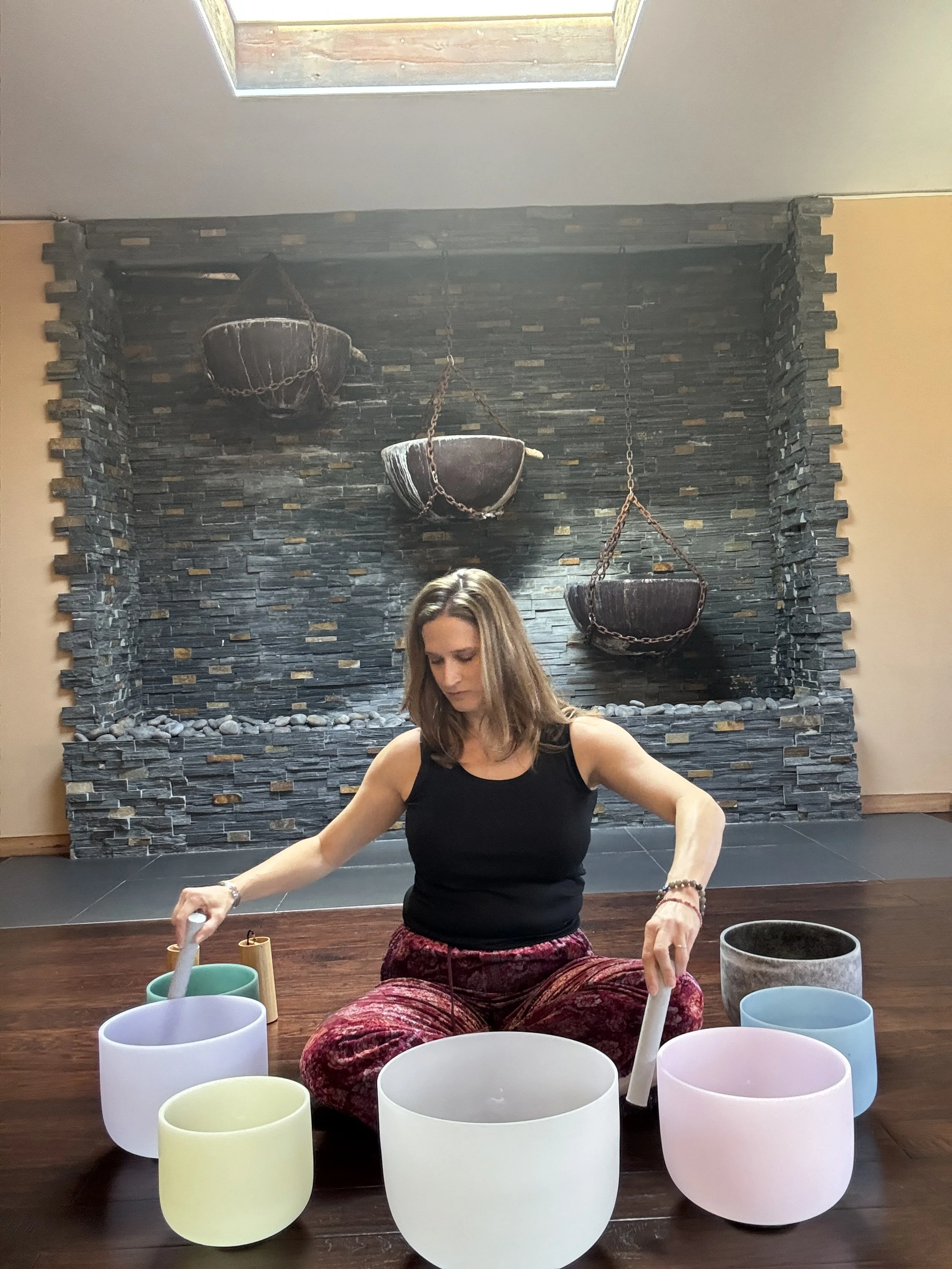 A woman sitting cross-legged on a wooden floor, playing crystal singing bowls with a mallet in each hand, in front of a textured stone wall with hanging hanging bowls in a wellness or meditation room.