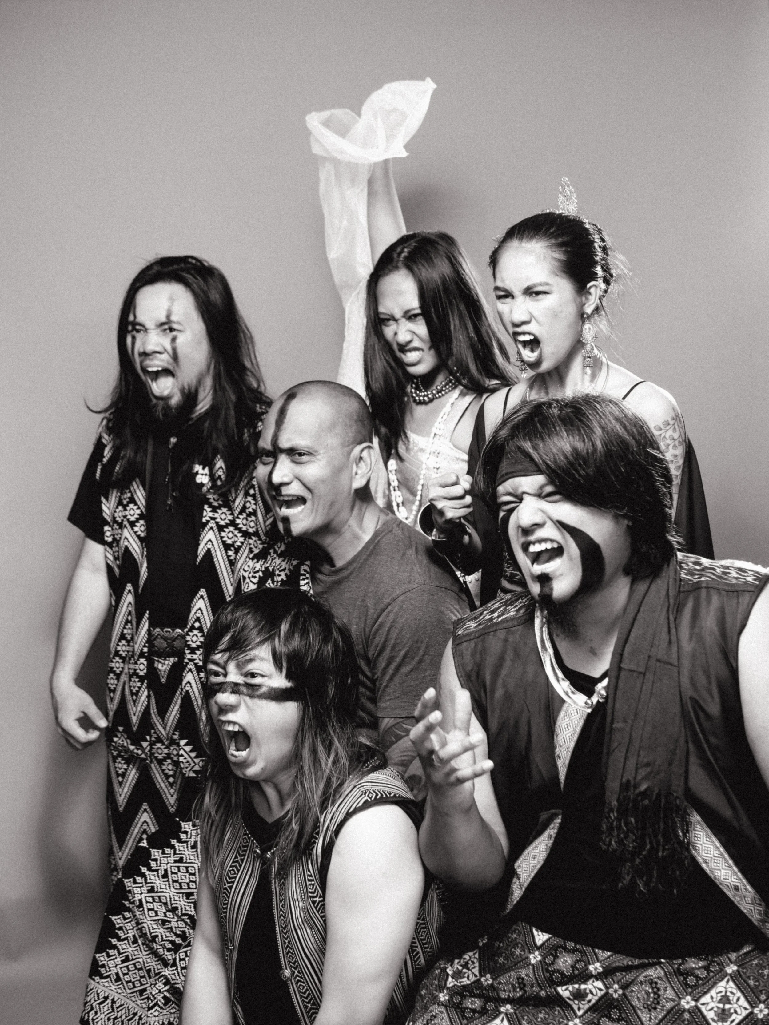 Group of six people, five women and one man, with intense facial expressions, shouting, and making gestures against a plain background. One woman is holding a plastic bag above her head.