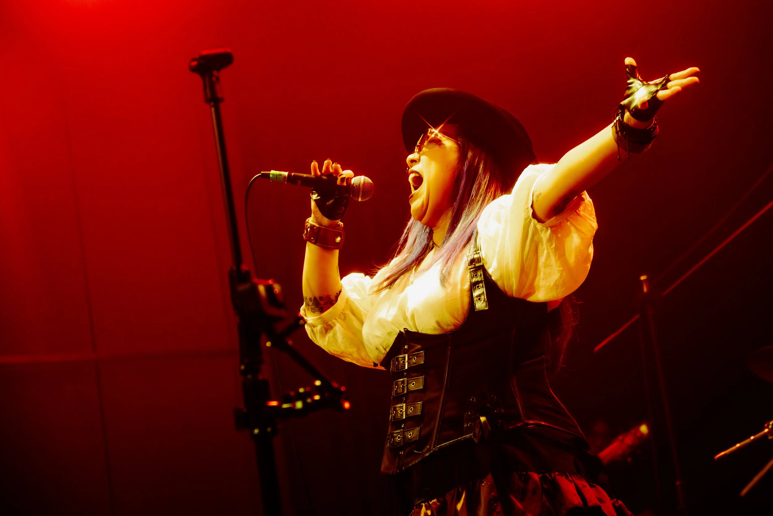 Performer singing passionately into a microphone on stage, wearing sunglasses, a black hat, and a black corset, with red stage lighting illuminating the scene.