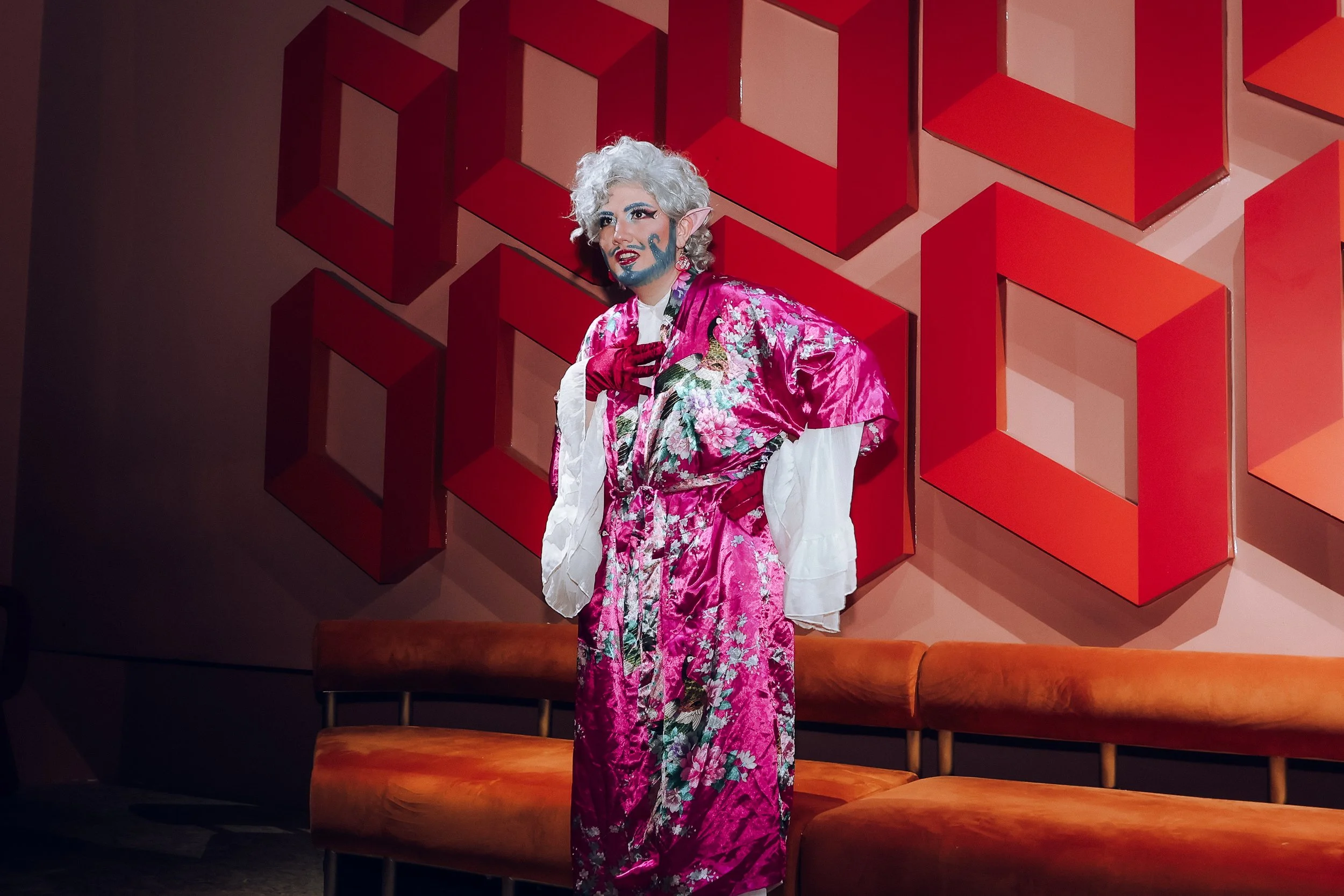 Person dressed in elaborate costume with makeup and wig standing in front of a geometric red art installation, with orange benches below.