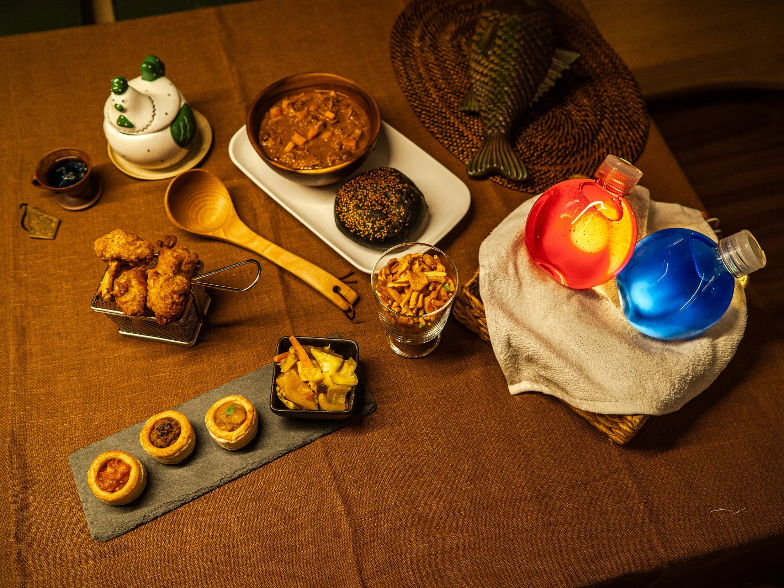 A variety of food items on a brown table, including a teapot with a bear design, a bowl of hearty stew, a black sesame covered bun, fried chicken, a glass of noodle salad, a slate platter of savory tarts, and colorful decorative lamps.