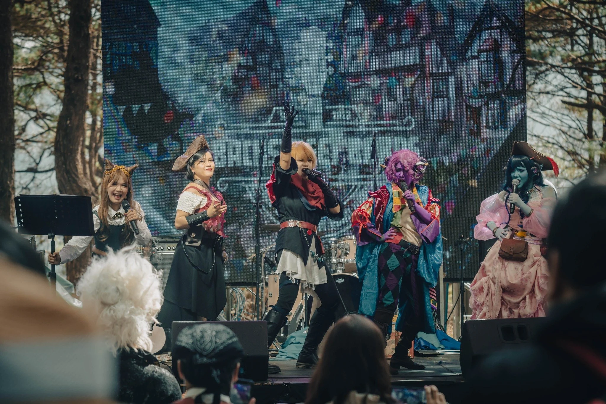 Cosplayers performing at an outdoor event with a large digital backdrop displaying a building and the words "Backstage Pass".