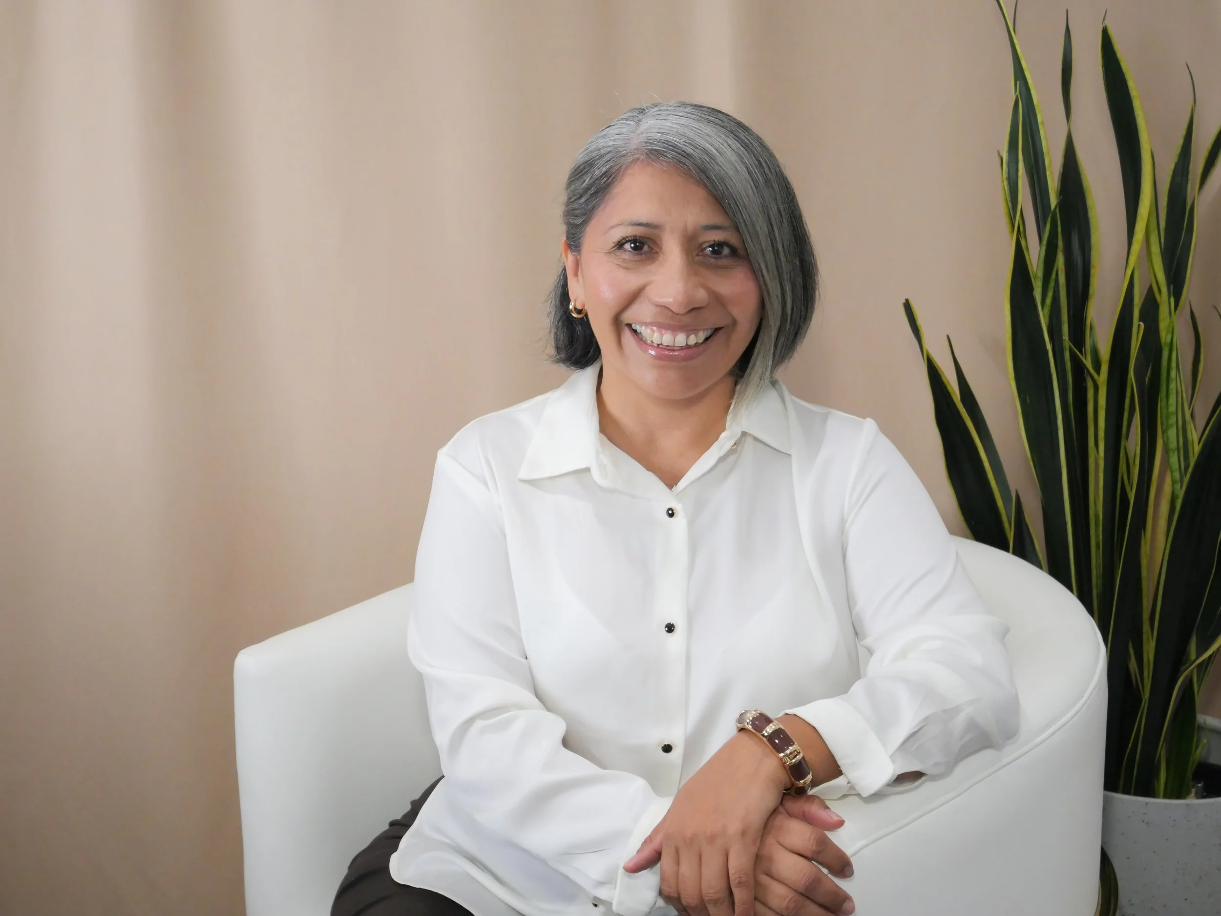 A smiling middle-aged woman with gray hair wearing a white button-down shirt, sitting on a white chair with her hands resting on her lap, and a tall green plant in the background.