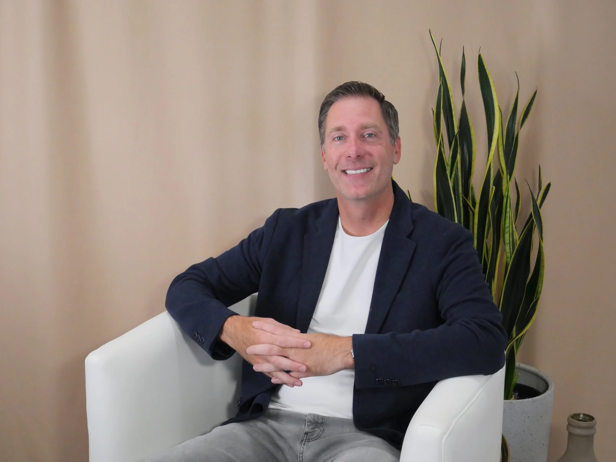 A smiling man with short brown hair, wearing a white t-shirt and navy blazer, seated on a white chair, with a tall green plant in a white pot behind him and a beige wall in the background.
