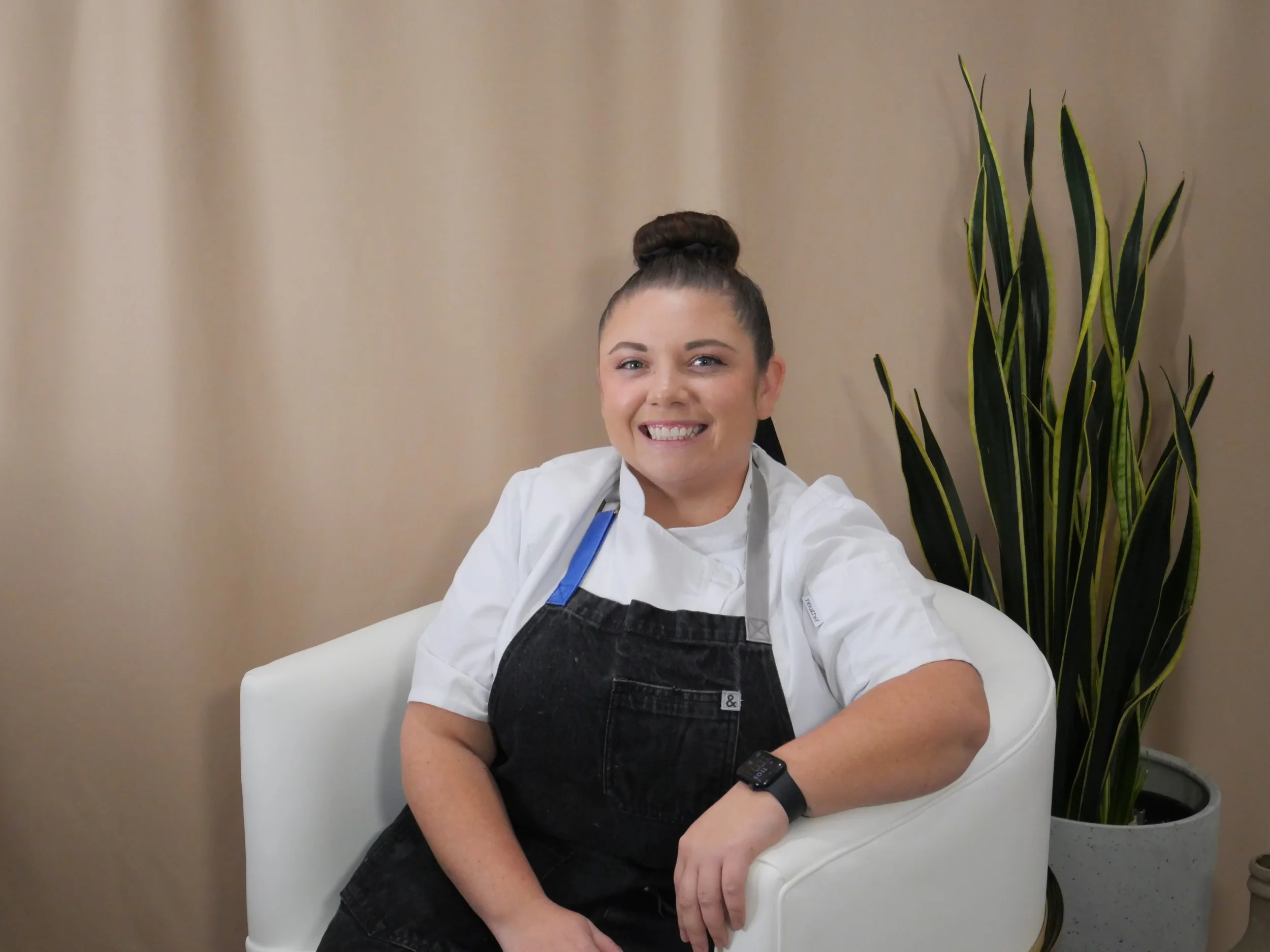 A smiling woman with dark hair in a bun, wearing a chef's coat and black apron, sitting on a white chair next to a tall green plant in a gray pot, in front of a beige curtain.
