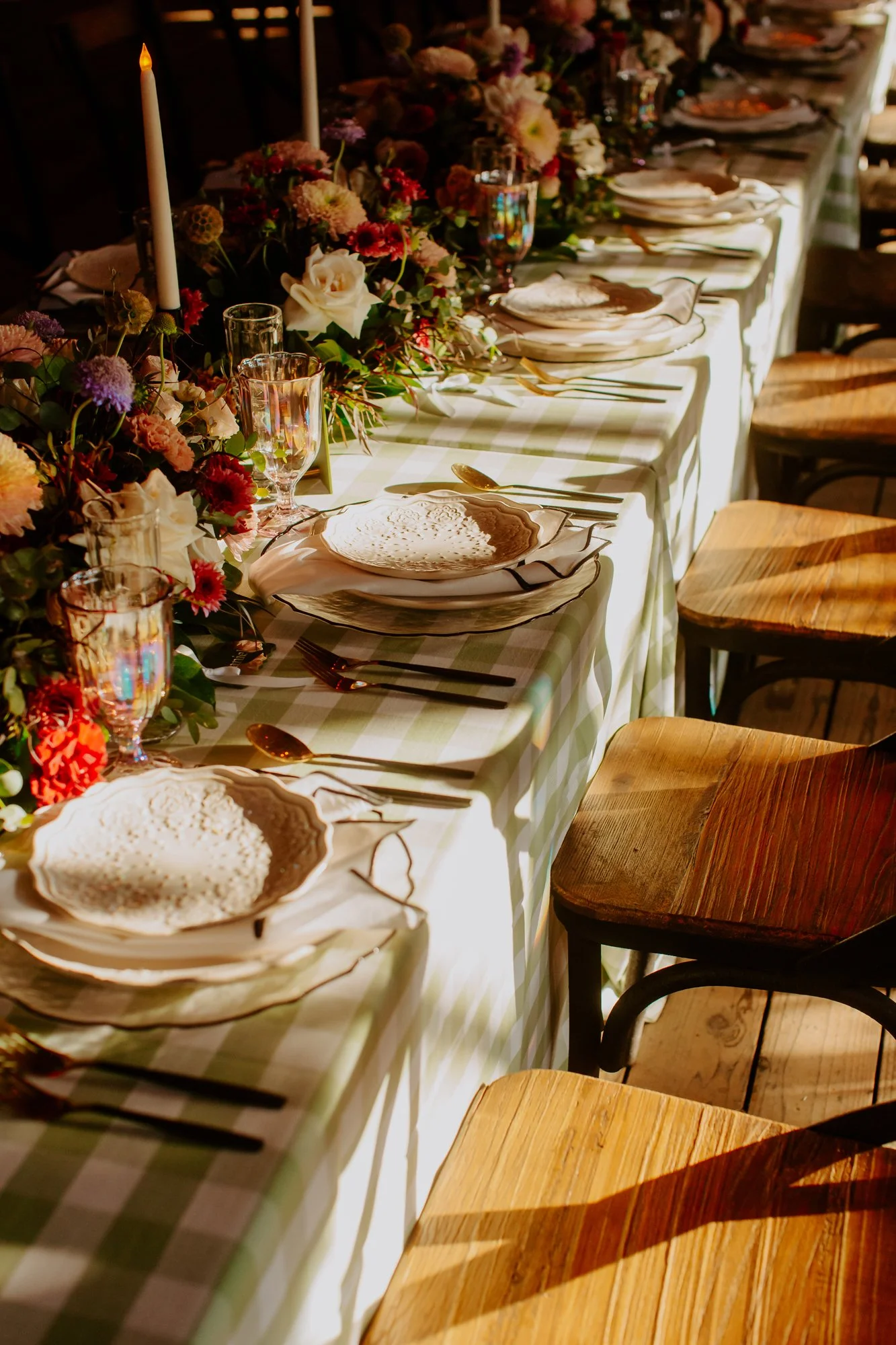 Elegant long dining table set with floral centerpiece, candles, fine china, glassware, and gold utensils, illuminated by warm sunlight.