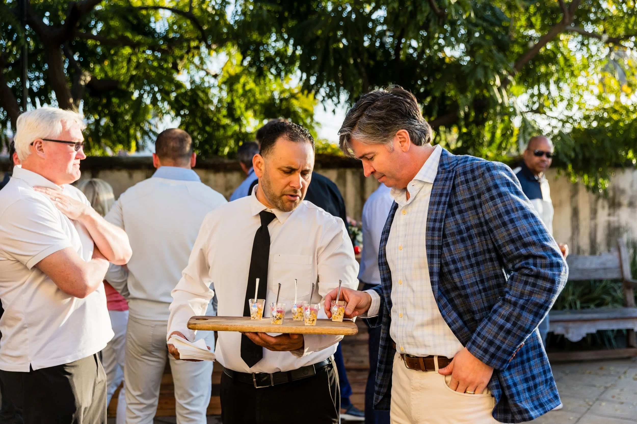 A man in a white shirt and black tie holding a tray of small dessert cups, serving a man in a blue checkered blazer at an outdoor gathering with other people in the background.