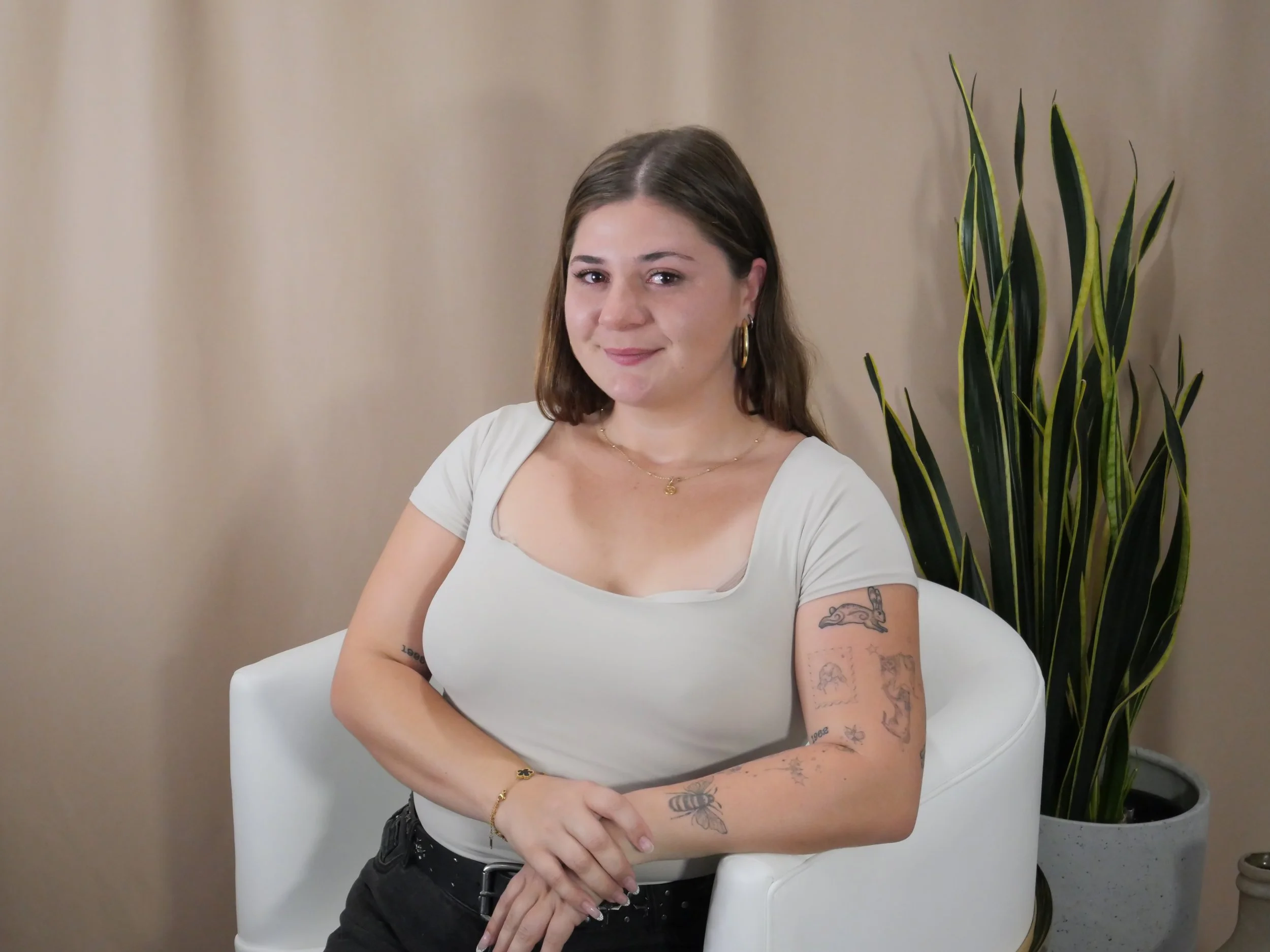 Young woman with brown hair, tattoos on her arms, wearing a white t-shirt, sitting in a white chair with a plant behind her.