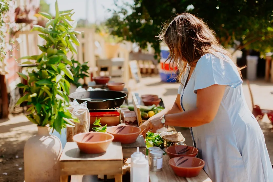 A woman in a white dress preparing food outdoors at a table with various bowls and plants.