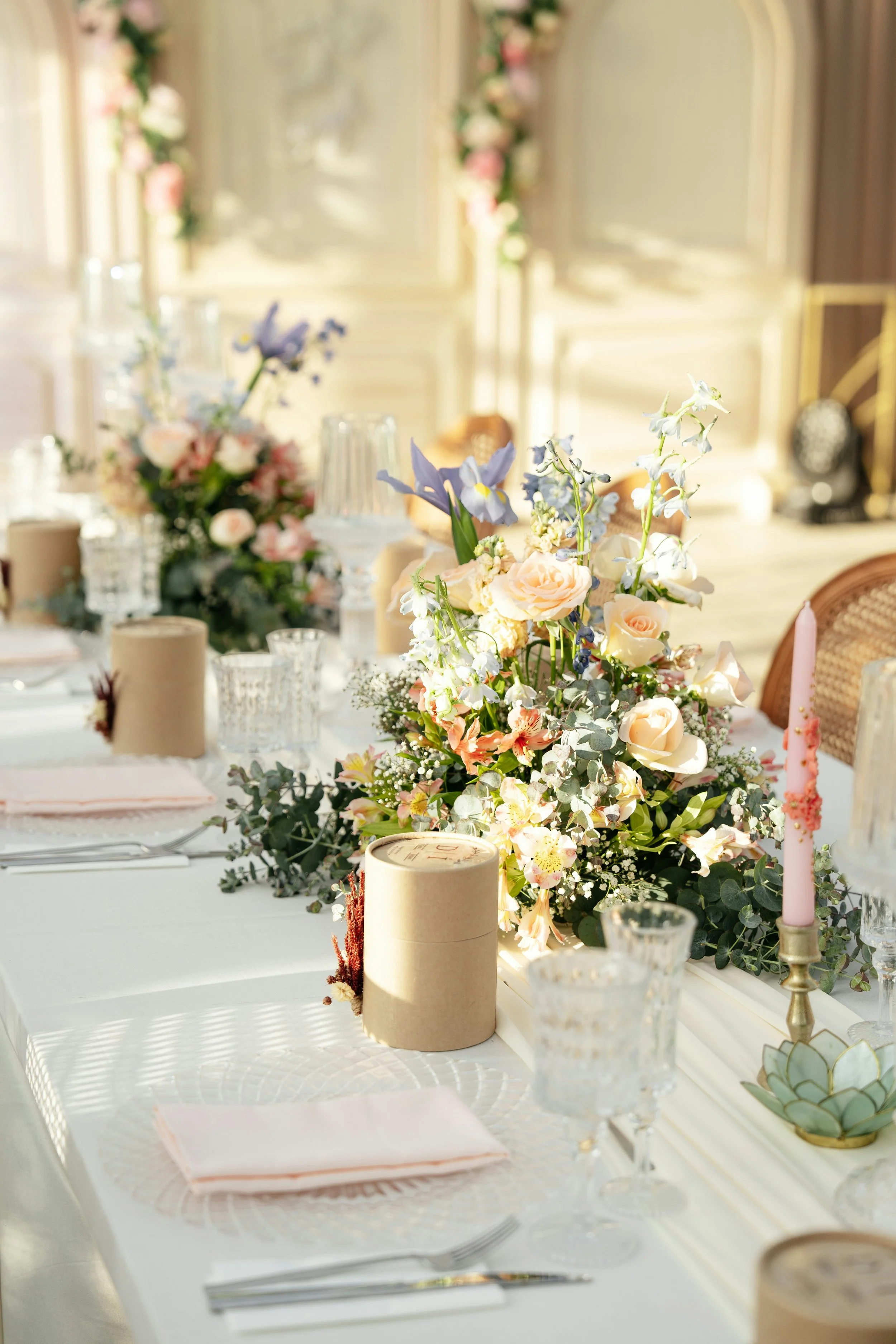 Elegant table decorated for a wedding or special event with a floral centerpiece, pink candles, glassware, and beige wrapped items.
