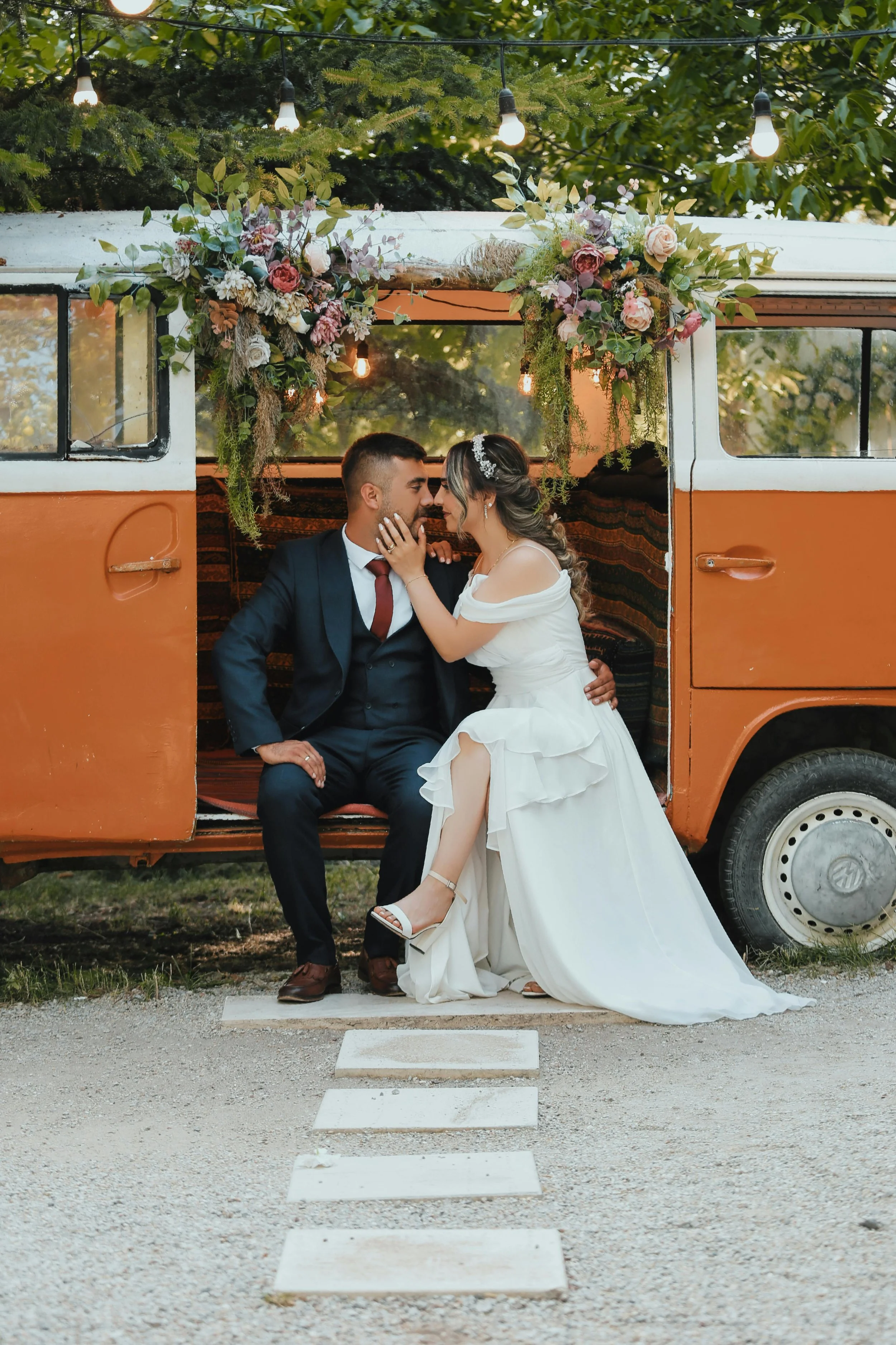 A couple in wedding attire sitting in an orange vintage van decorated with flowers and greenery, outside surrounded by trees, during a wedding photoshoot.