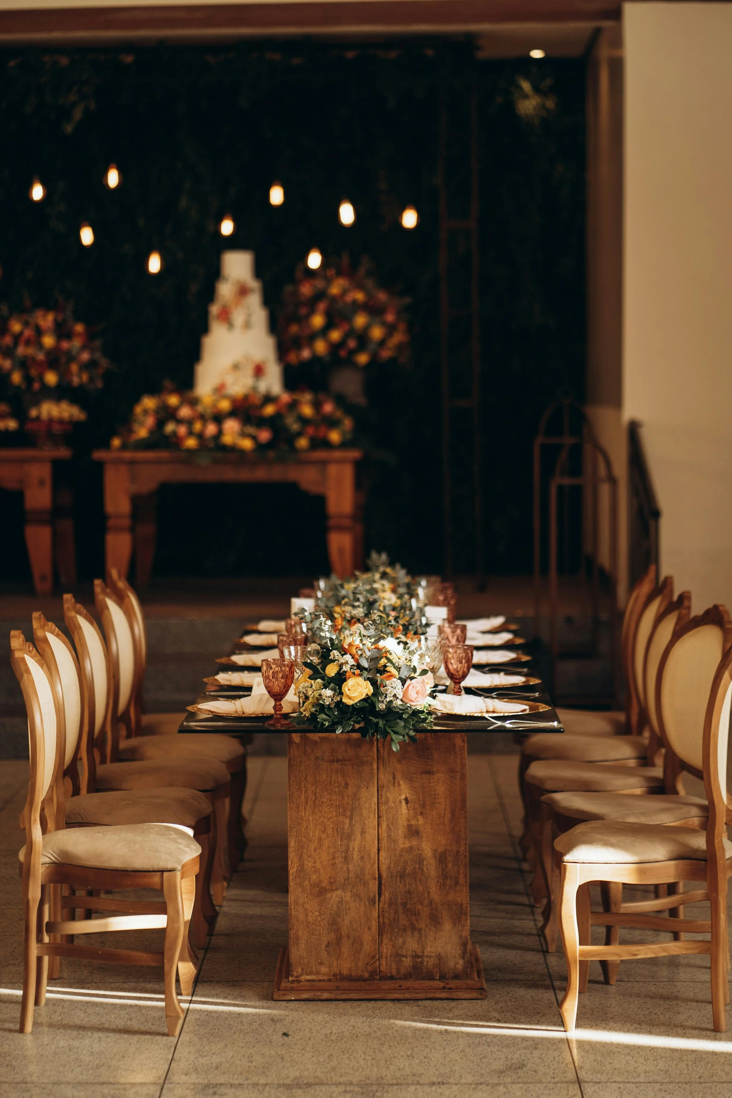 Elegant wedding reception table with a floral centerpiece, set with napkins, glasses, and plates, in front of a cake and floral arrangements on a dark background.