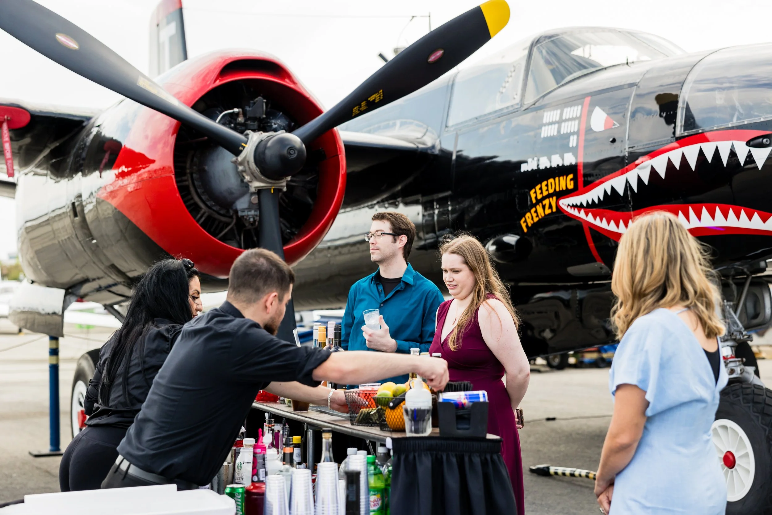 Group of people gathered around a table of drinks and snacks in front of a vintage airplane, which has a shark mouth painted on its nose and the words 'Feeding Frenzy' on its side.
