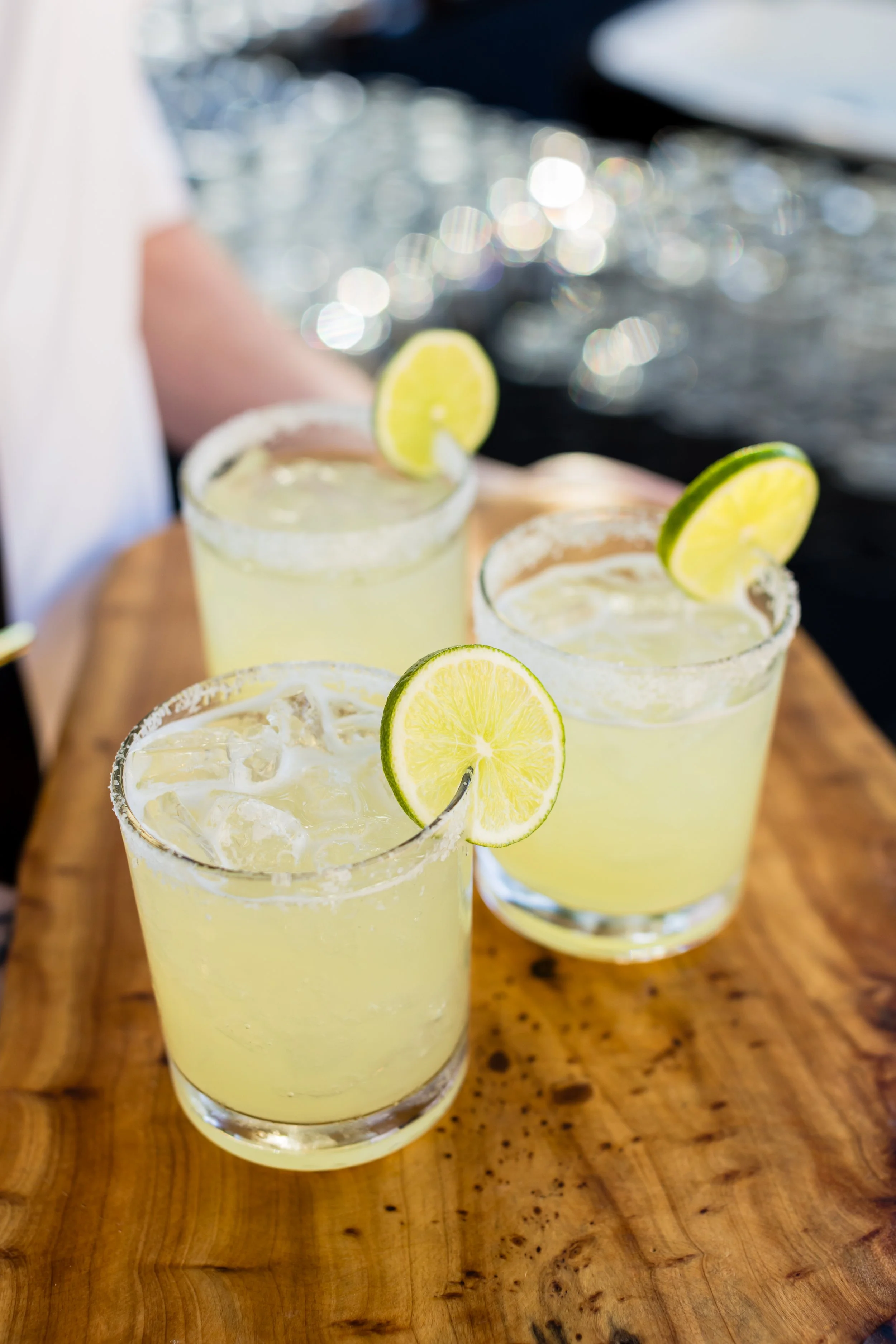 Three glasses of margaritas with salt-rimmed edges, garnished with lime and lemon wedges, on a wooden serving tray.