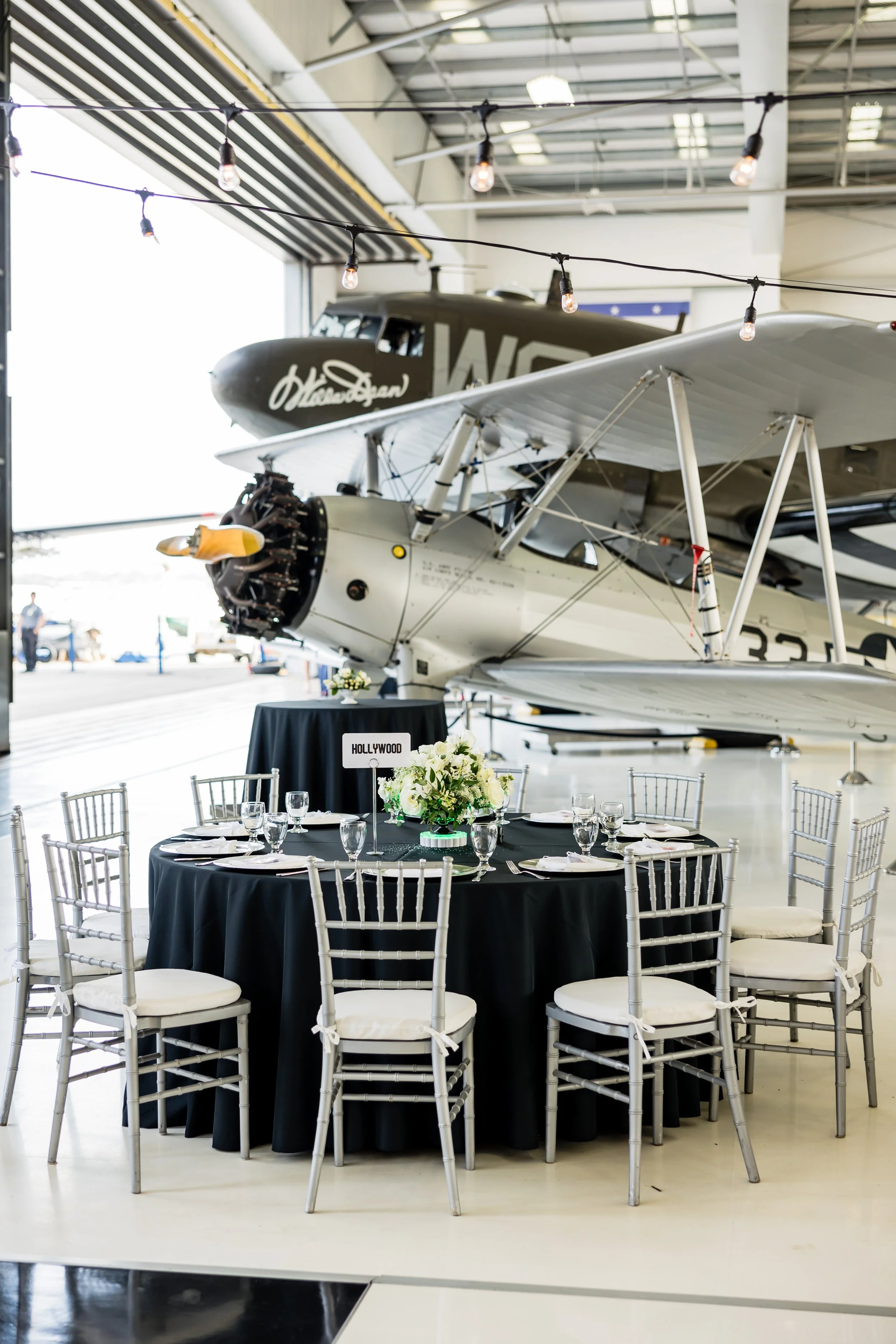 Event setup with a round table covered with a black tablecloth, silver chairs, floral centerpiece, set for a meal, situated in front of vintage aircraft inside an airplane hangar.