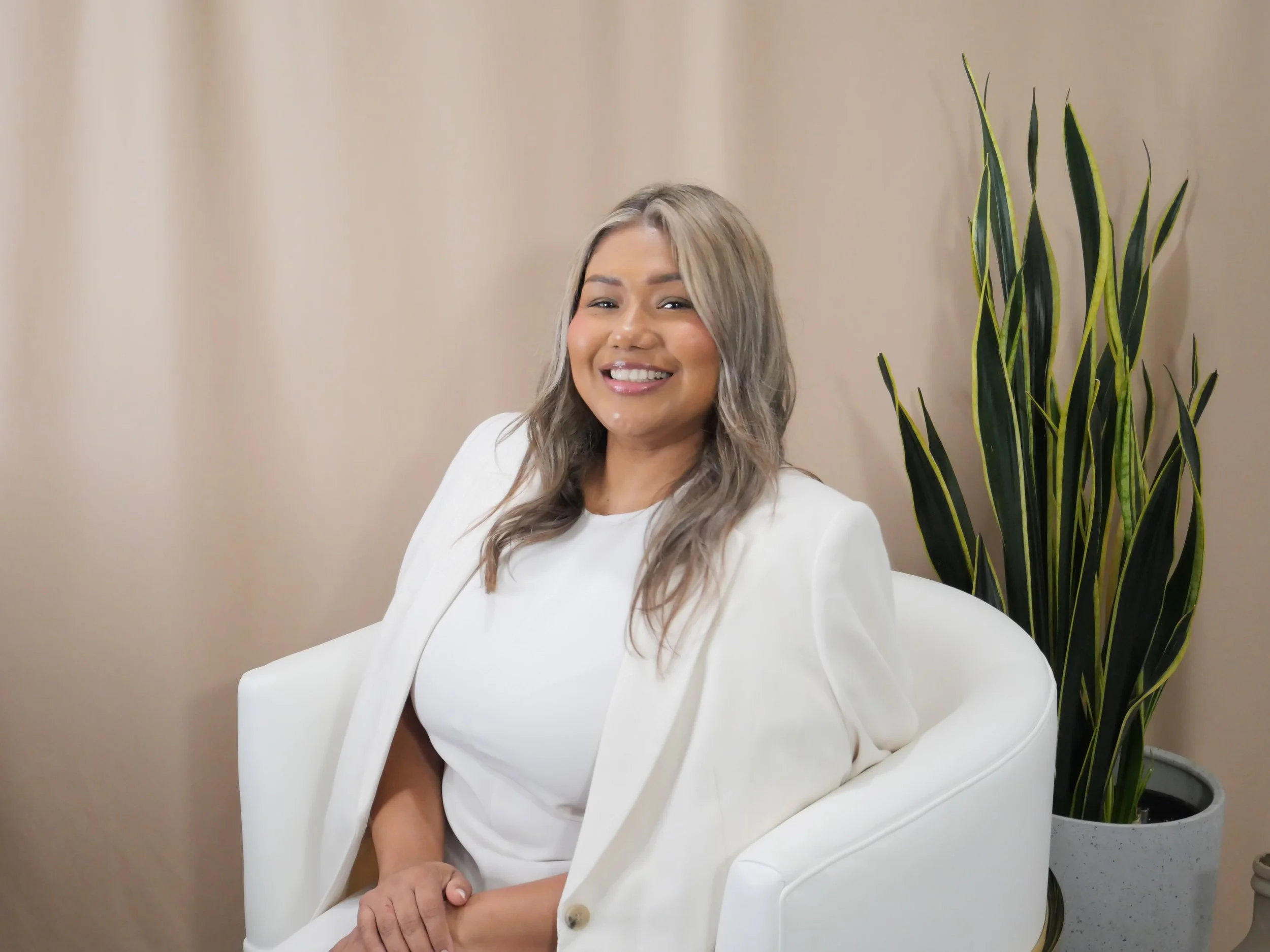 A woman with wavy blonde hair smiling and sitting in a white armchair, wearing a white blazer over a white top, with a large green potted plant behind her against a beige background.