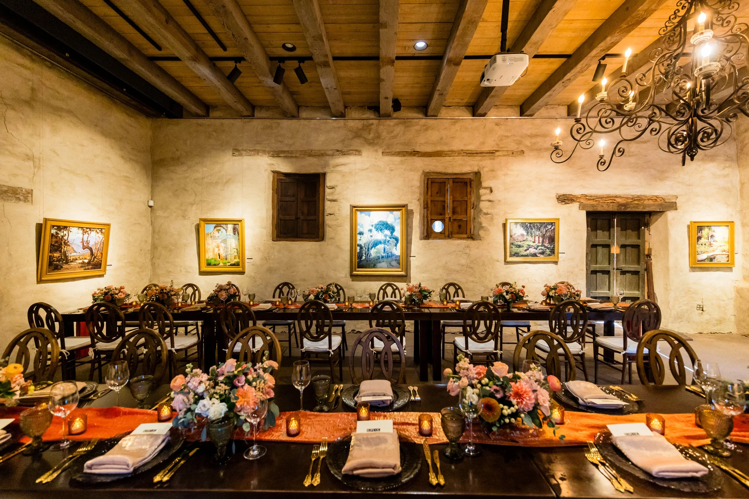 Interior of a rustic dining room decorated for an event, featuring long tables with floral centerpieces, candles, and elegant place settings. The room has exposed wooden beams and framed artwork on textured walls, with vintage windows and a chandelier.