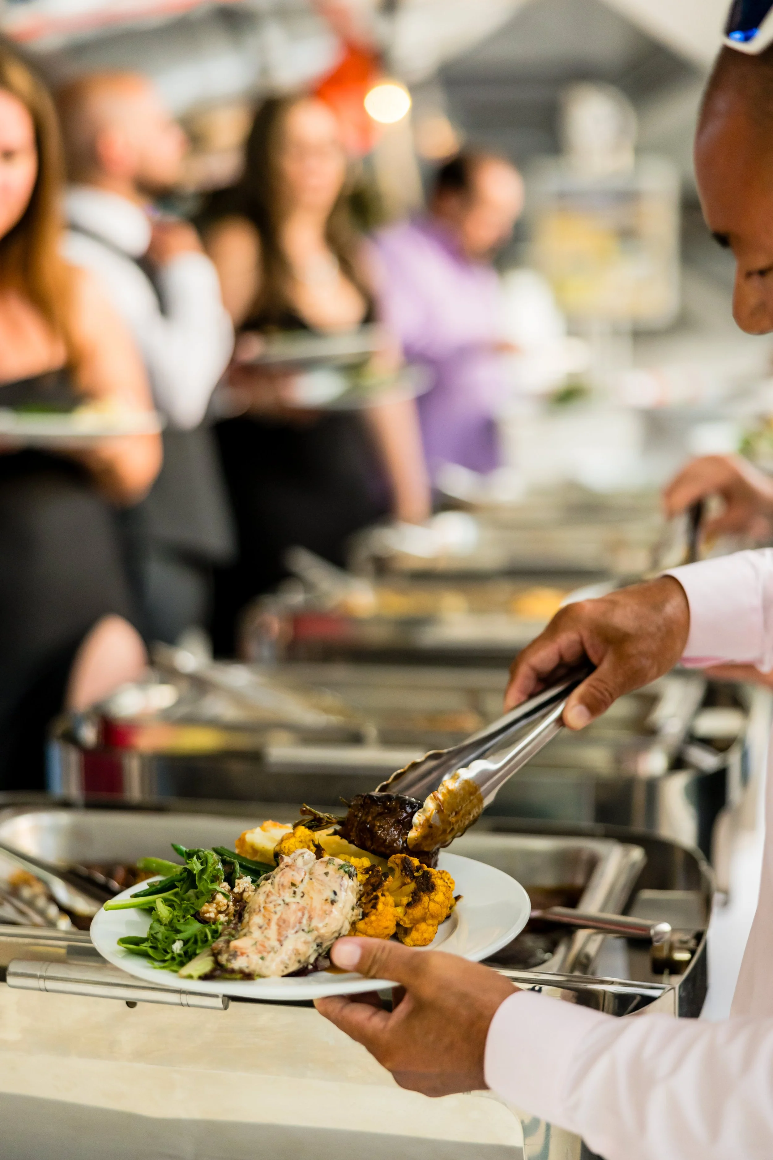 A person serving food at a buffet with other people waiting in line in the background.