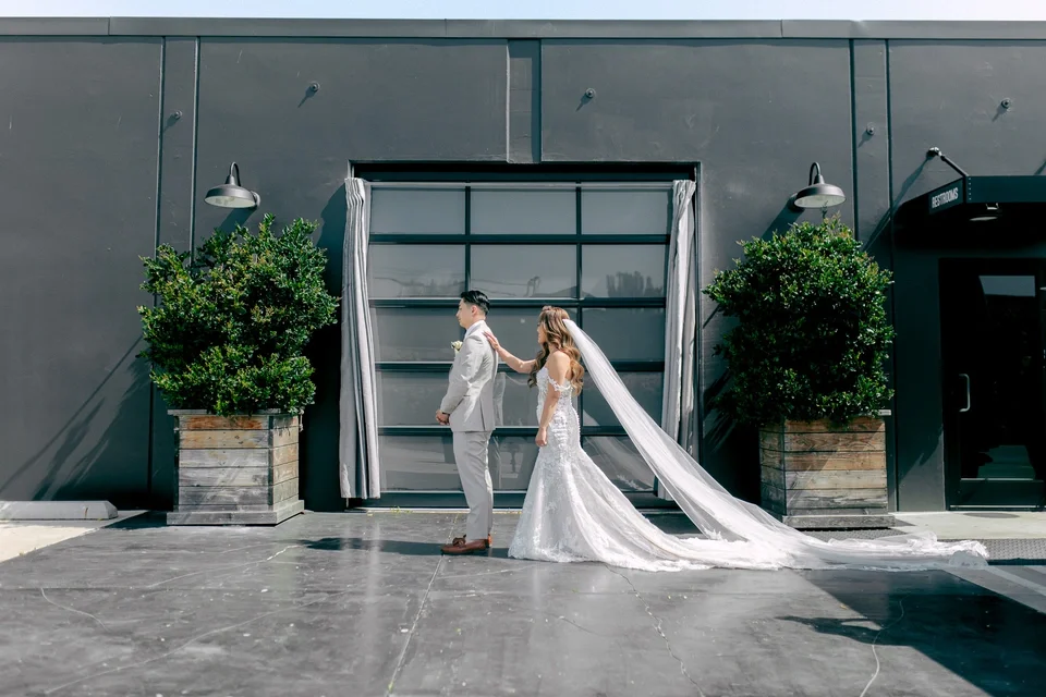 Bride and groom standing outside a modern building, with the bride gently pulling the groom's tie, both dressed in wedding attire.