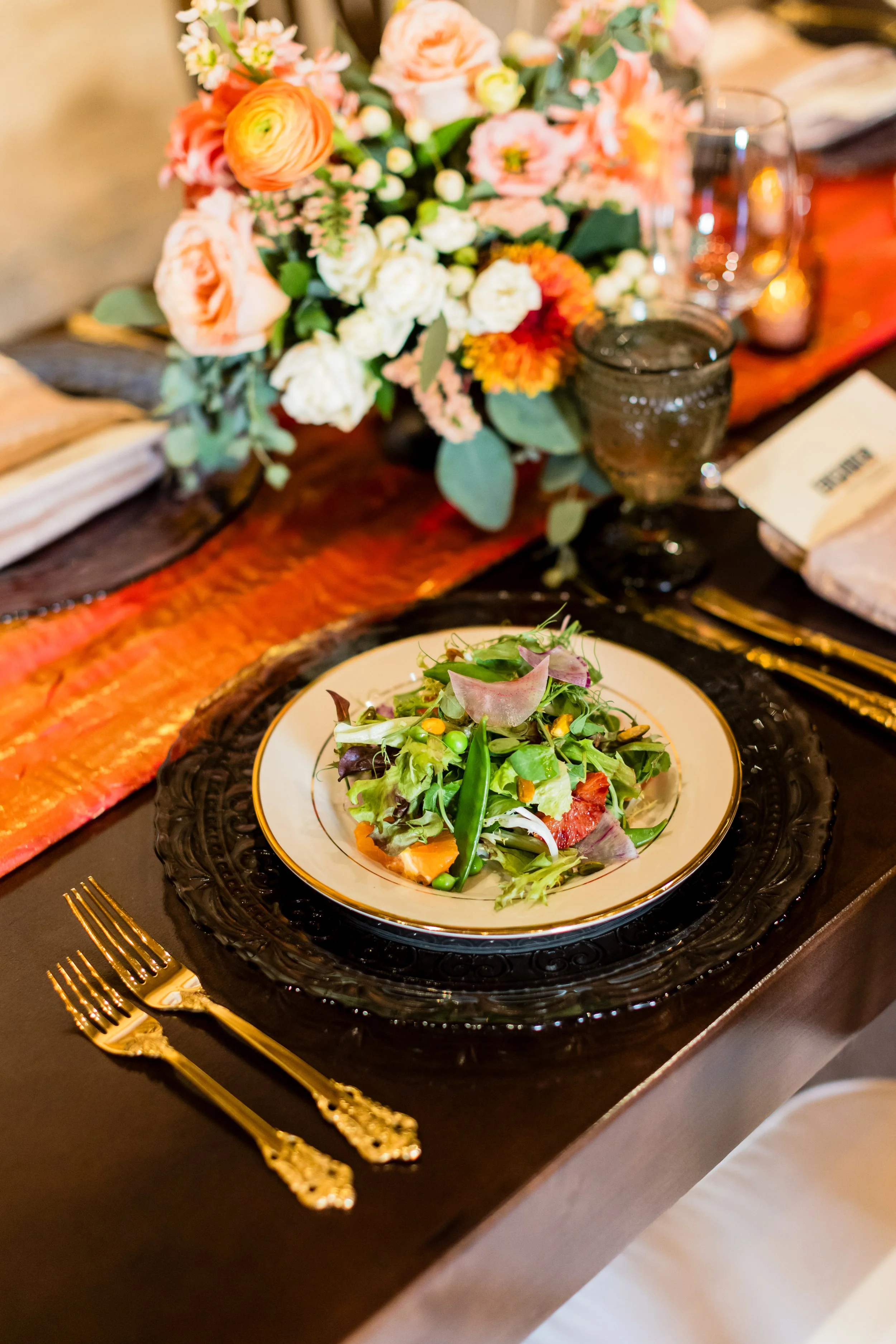 A floral centerpiece with pink, peach, white, and orange flowers on a rustic wooden table. A salad with mixed greens and vegetables is on a decorated plate, accompanied by gold utensils.
