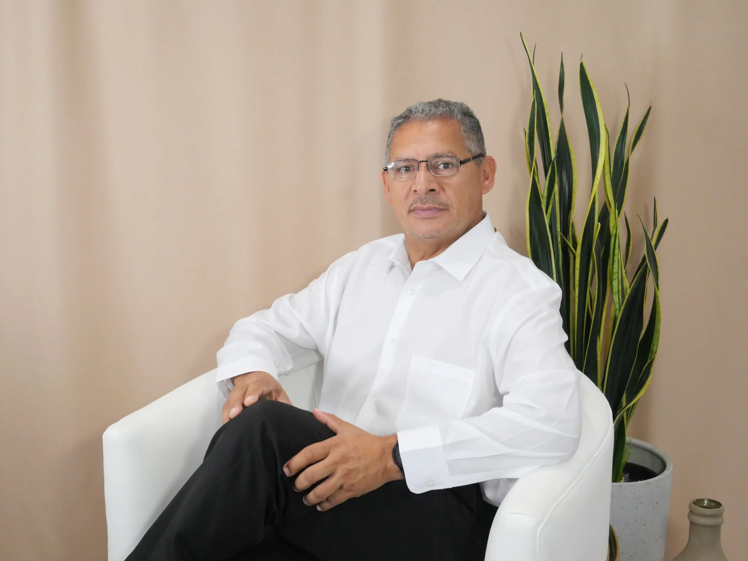 a middle-aged man with glasses, wearing a white shirt and black pants, sitting on a white chair, with a tall green plant in a pot behind him, against a beige background