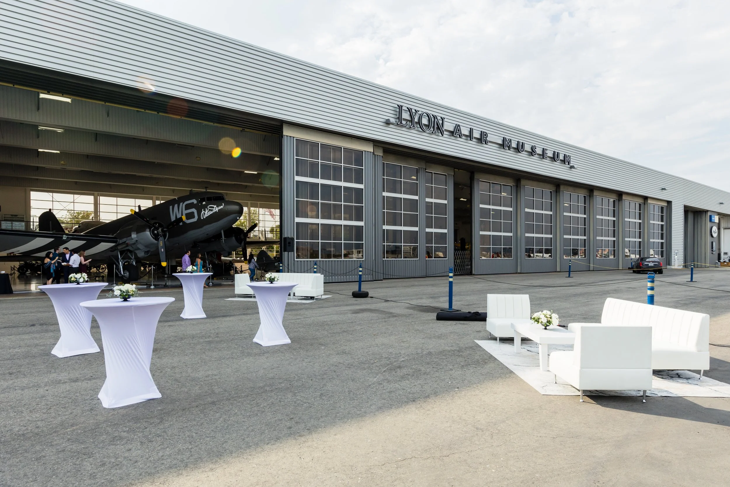 Outdoor view of Lyon Air Museum hangar with vintage airplane, white tables with flowers, and white seating area, in front of a large industrial-style building.