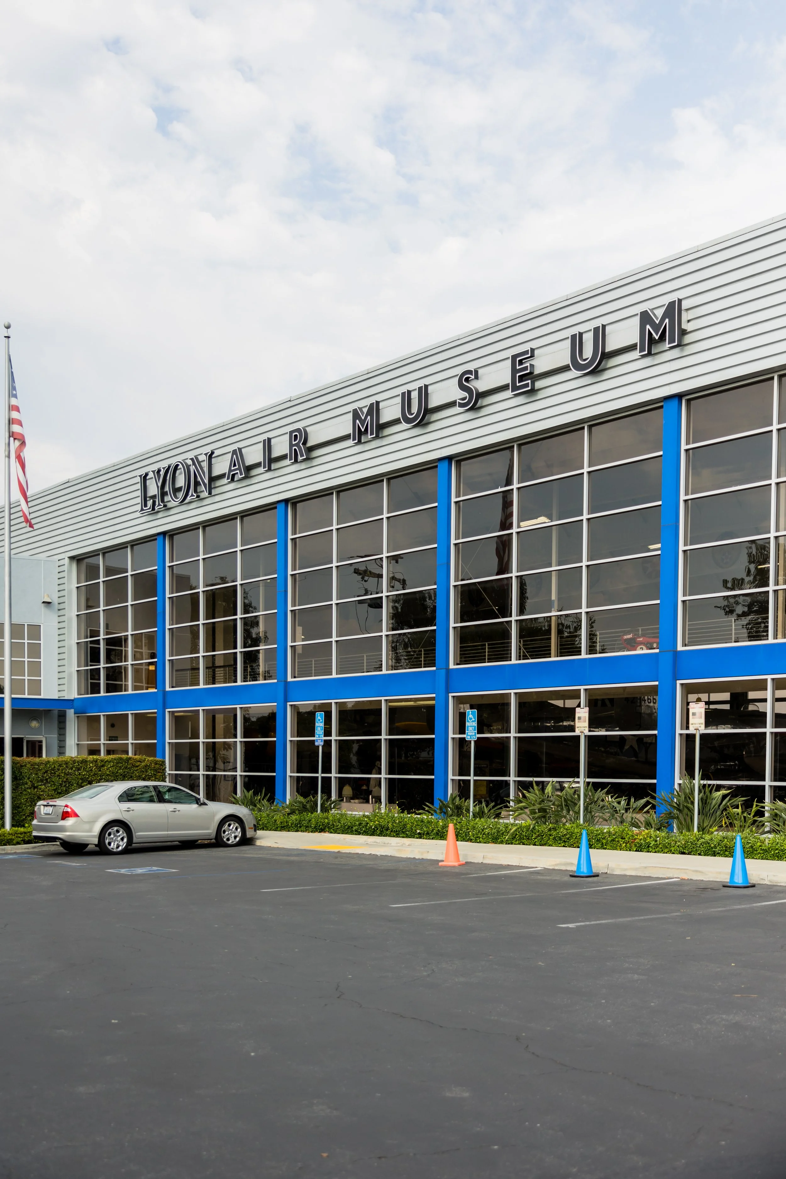 Exterior of Lyon Air Museum building with large glass windows, parking lot with a silver car, and colored cones, with the name 'Lyon Air Museum' on the facade.