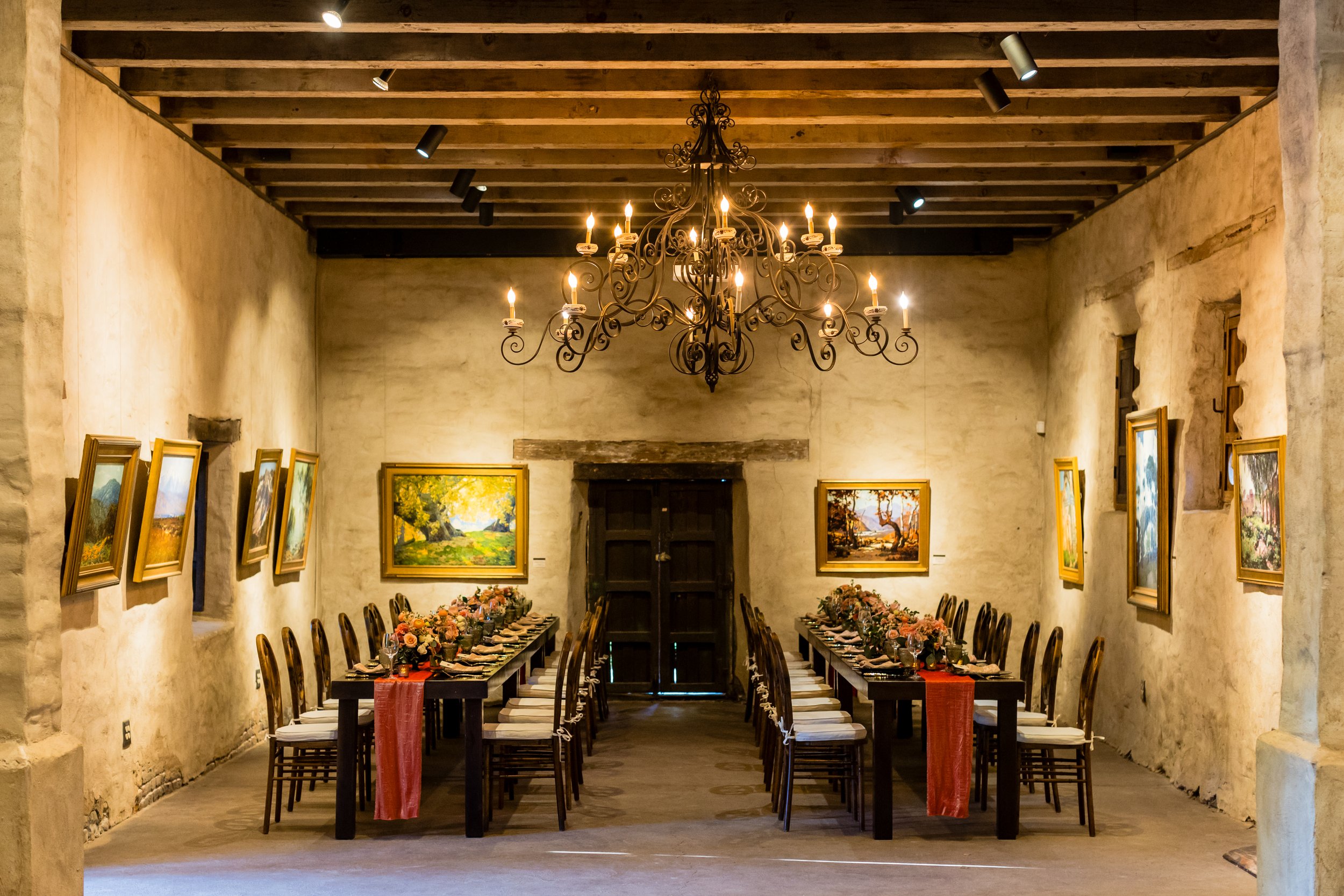 A rustic dining room with a chandelier, decorated for a special occasion, with two long tables set with floral centerpieces, plates, and cutlery. The room has stone walls, framed paintings, and a wooden ceiling.