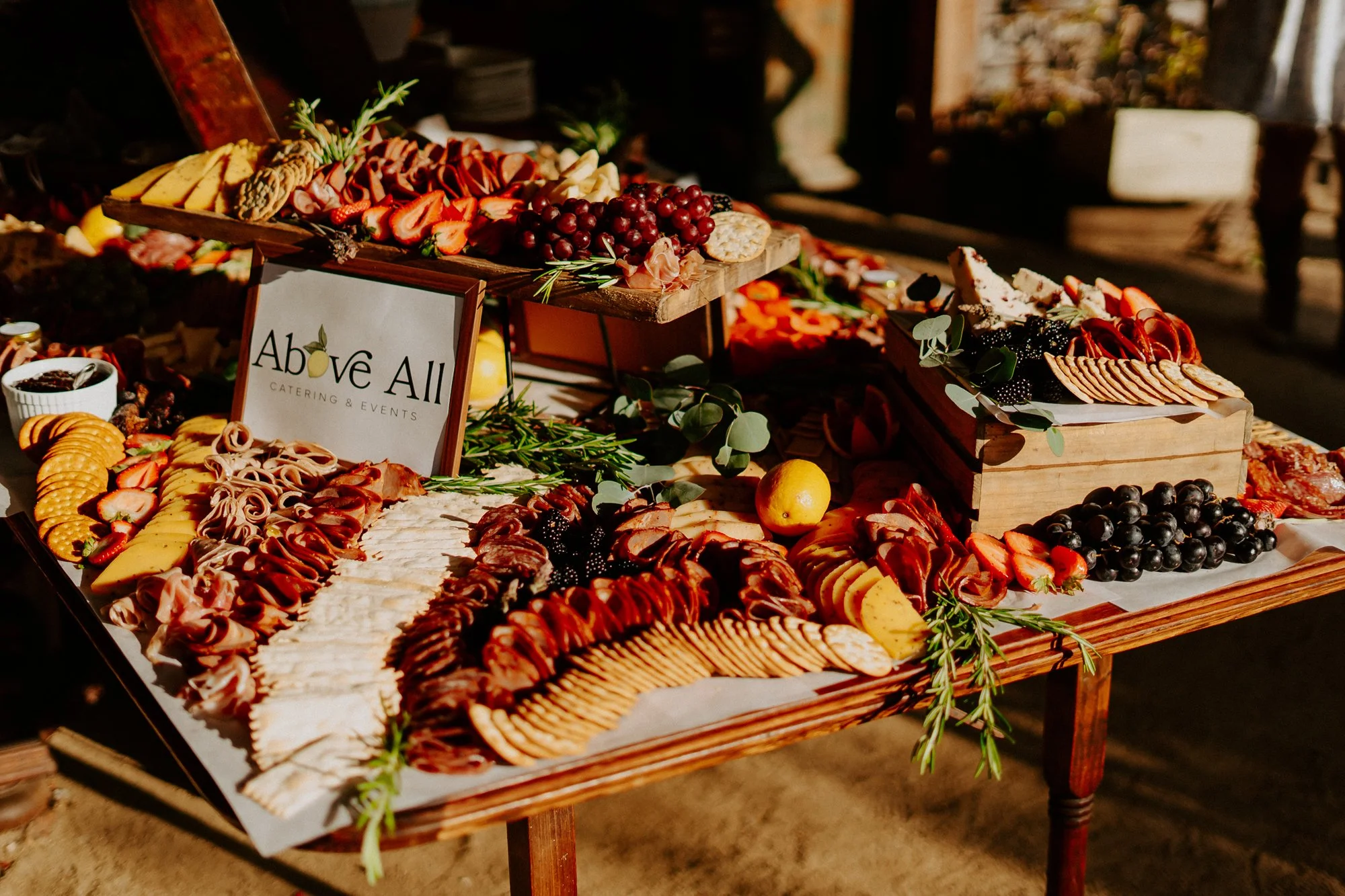 A wooden table displaying a variety of cheeses, meats, grapes, strawberries, and crackers, with a sign reading 'Above All Catering & Events' in the center.